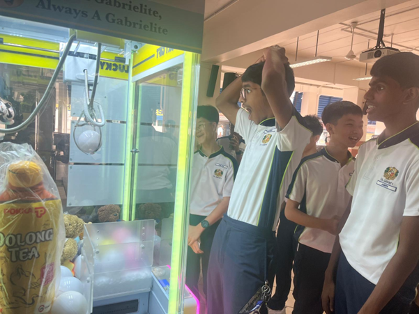 Several school students in uniforms watch and react excitedly near a claw machine filled with plush toys and drinks.