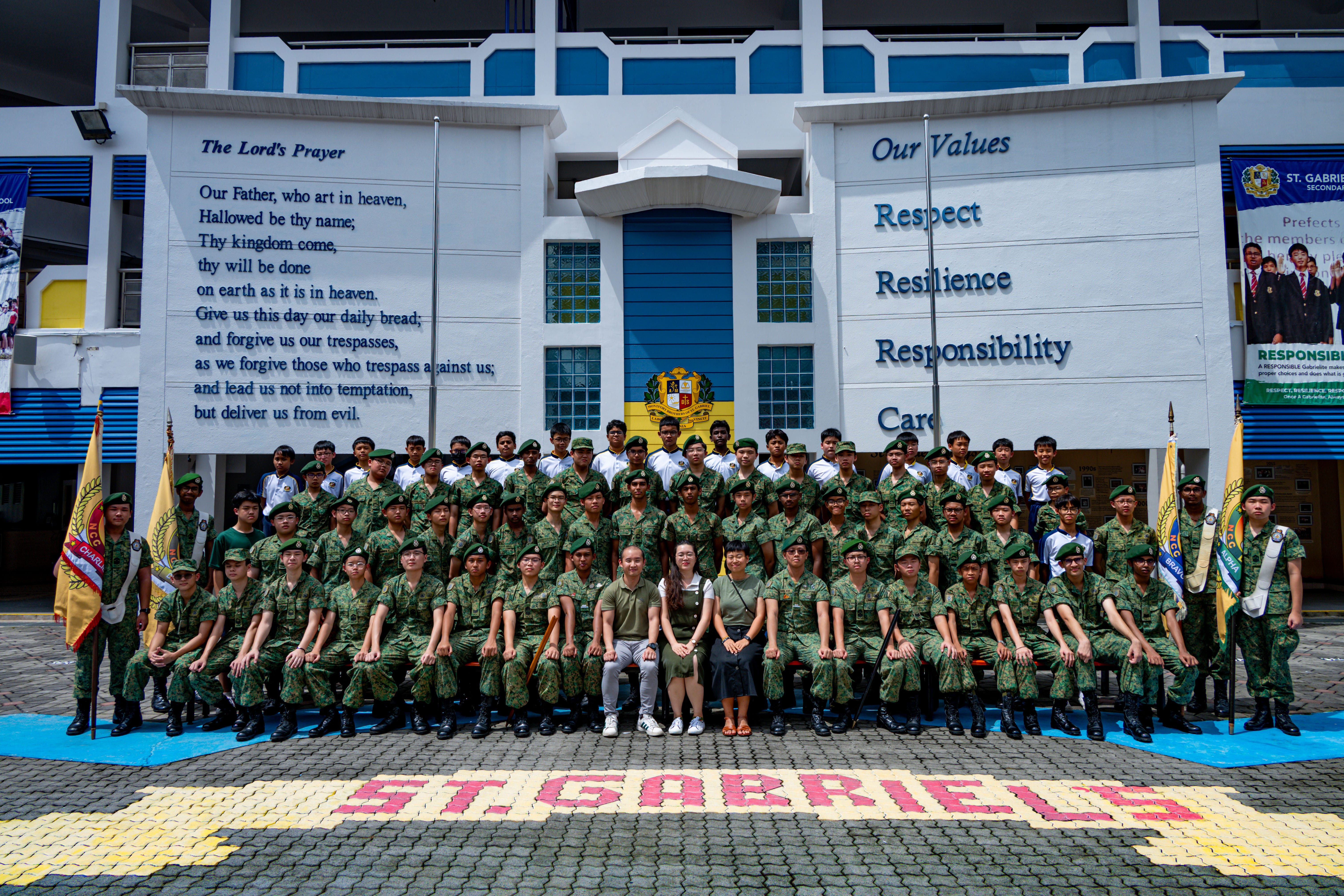 Large group in green uniforms posing at a school entrance with wall text The Lord's Prayer and Our Values.