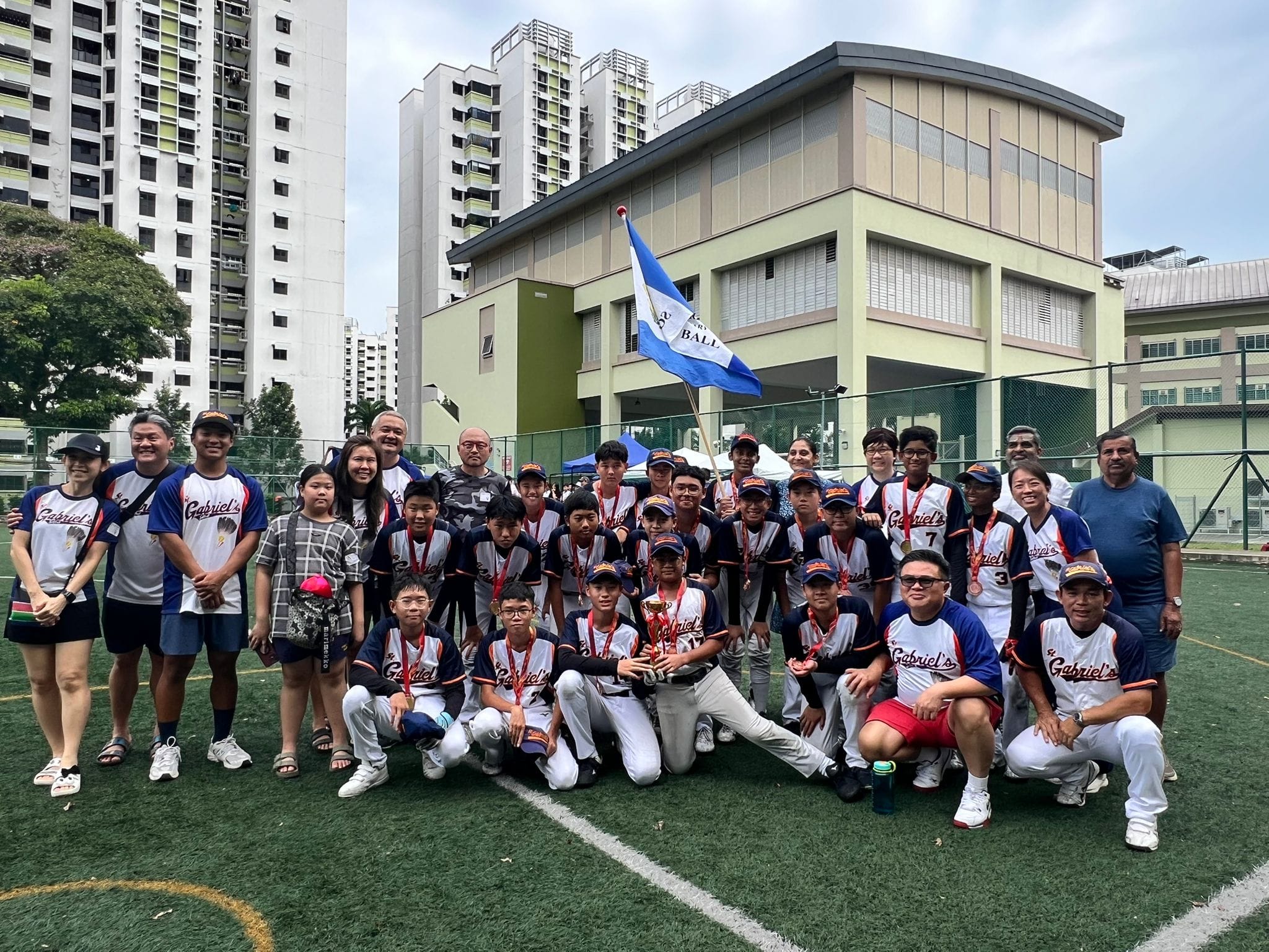 A youth baseball team in St. Gabriel's uniforms poses with a trophy and flag on a field near urban buildings.