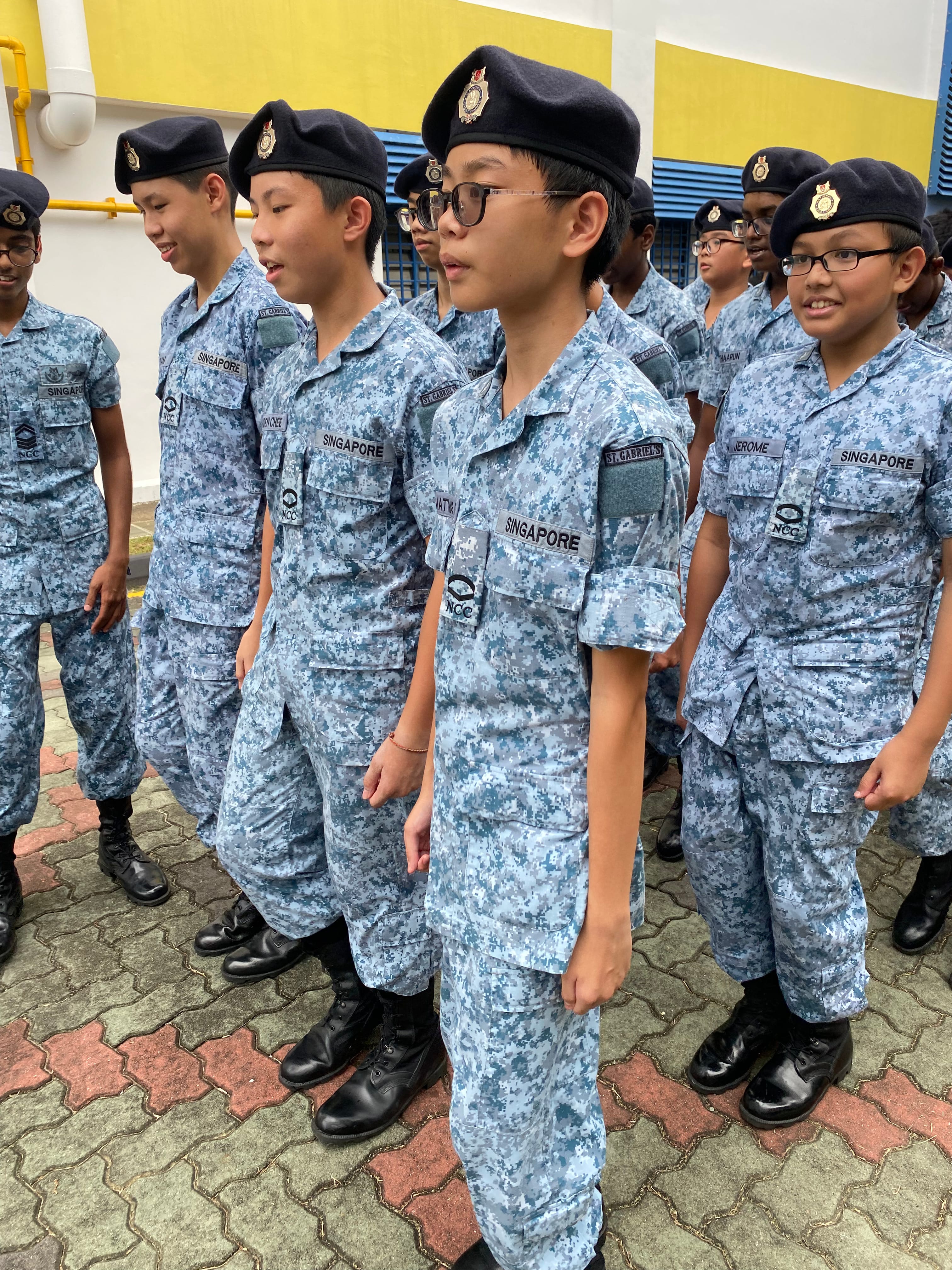 Group of youths in blue camouflage uniforms and black berets standing outdoors near a yellow wall