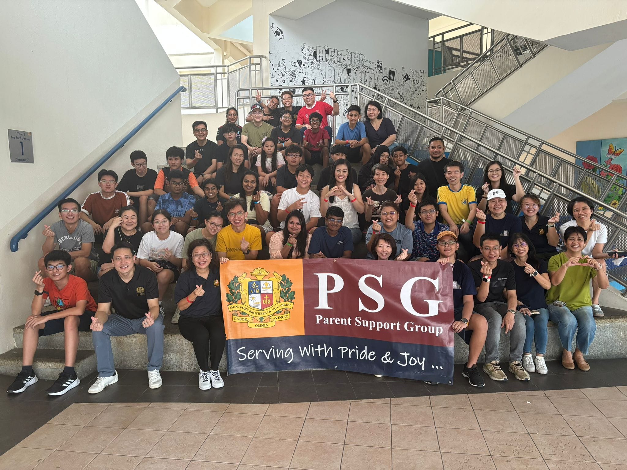 Large group of people sitting on indoor stairs holding a PSG Parent Support Group banner reading Serving With Pride and Joy.