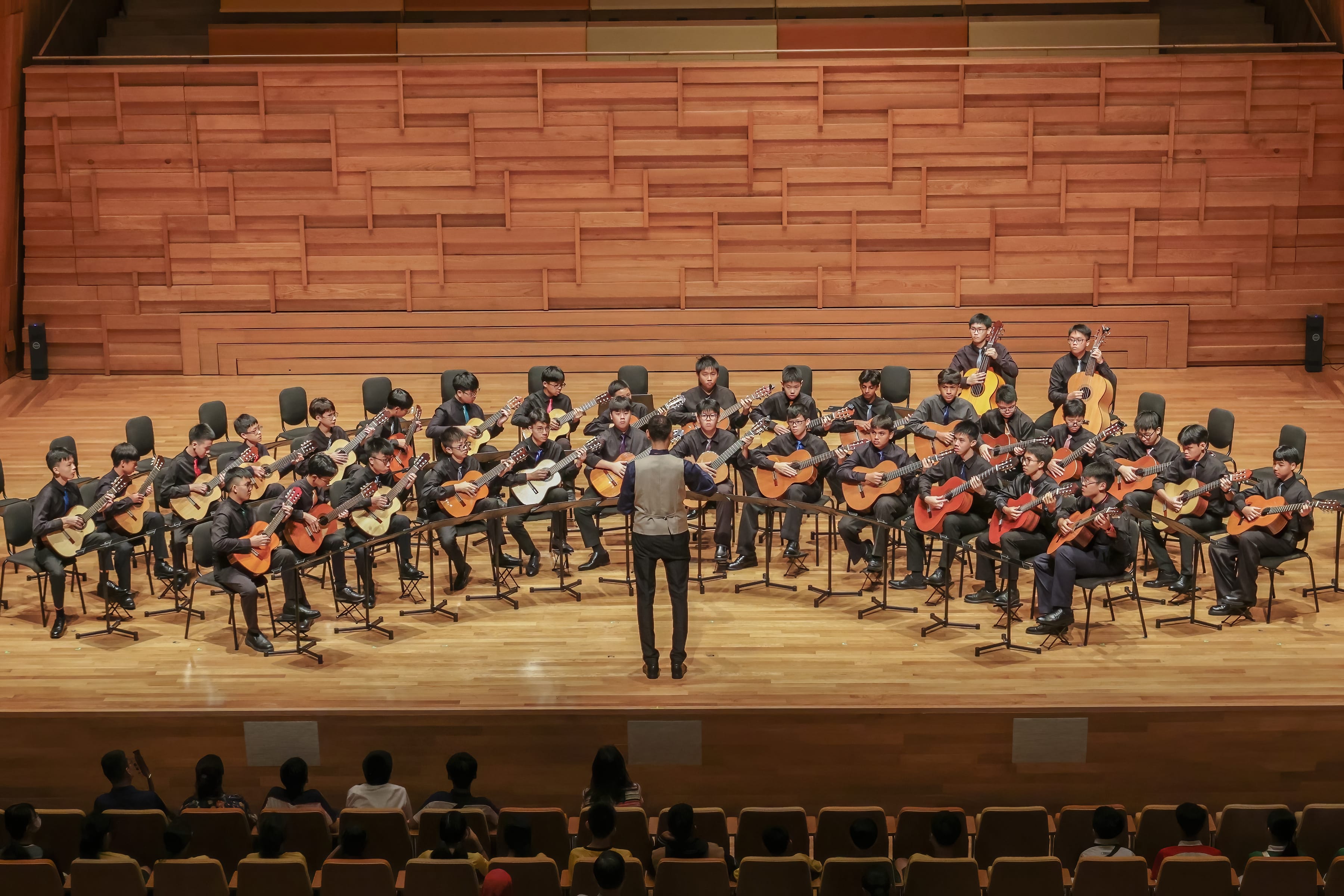 A conductor stands before a large youth guitar ensemble performing on a wooden concert stage before a seated audience.