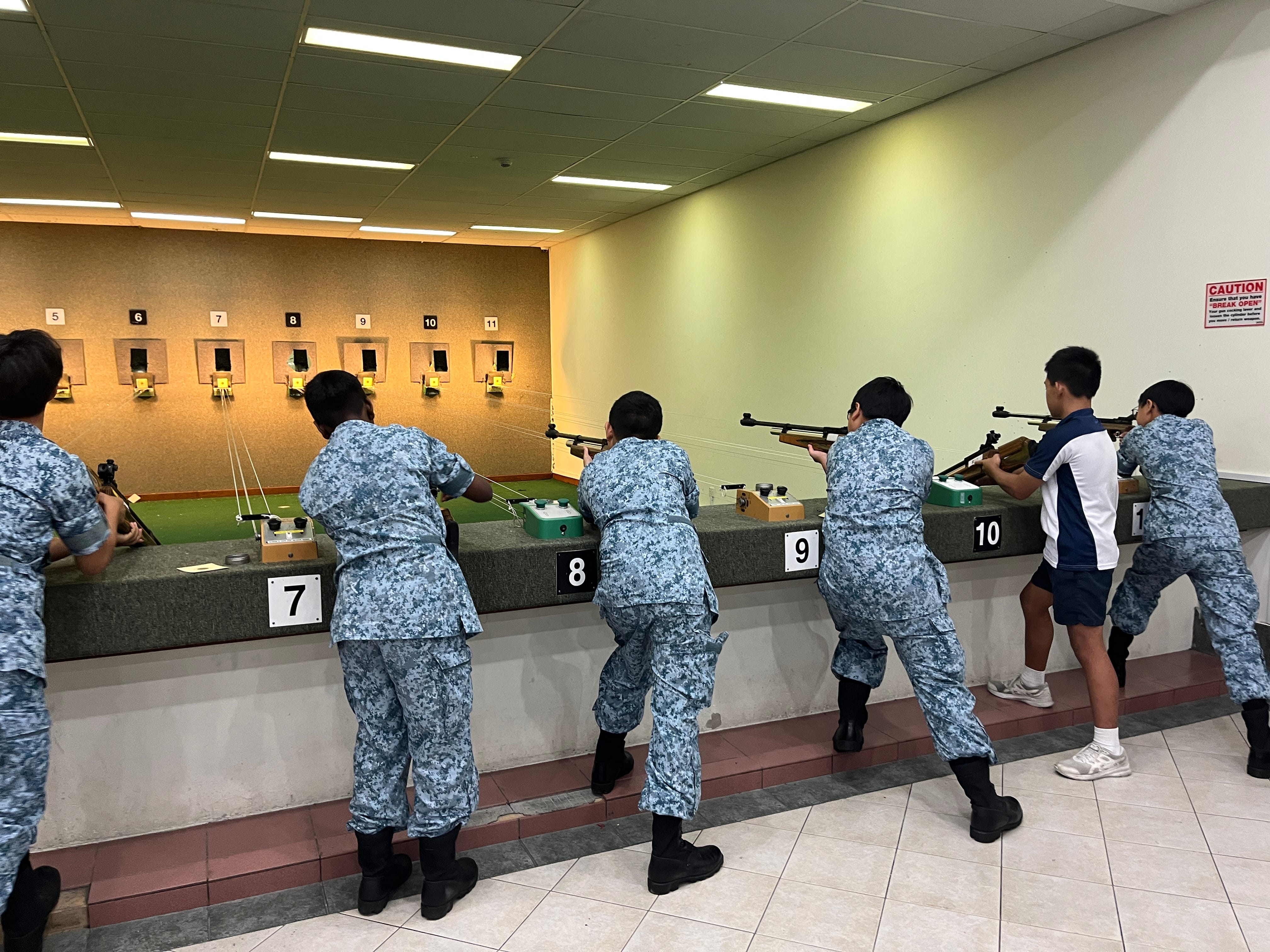 People in blue camouflage and one in sportswear aim rifles at numbered targets in an indoor range.
