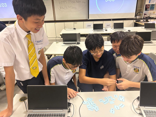 Five school boys in uniforms huddle around a desk with a laptop and scattered paper cutouts in a classroom.