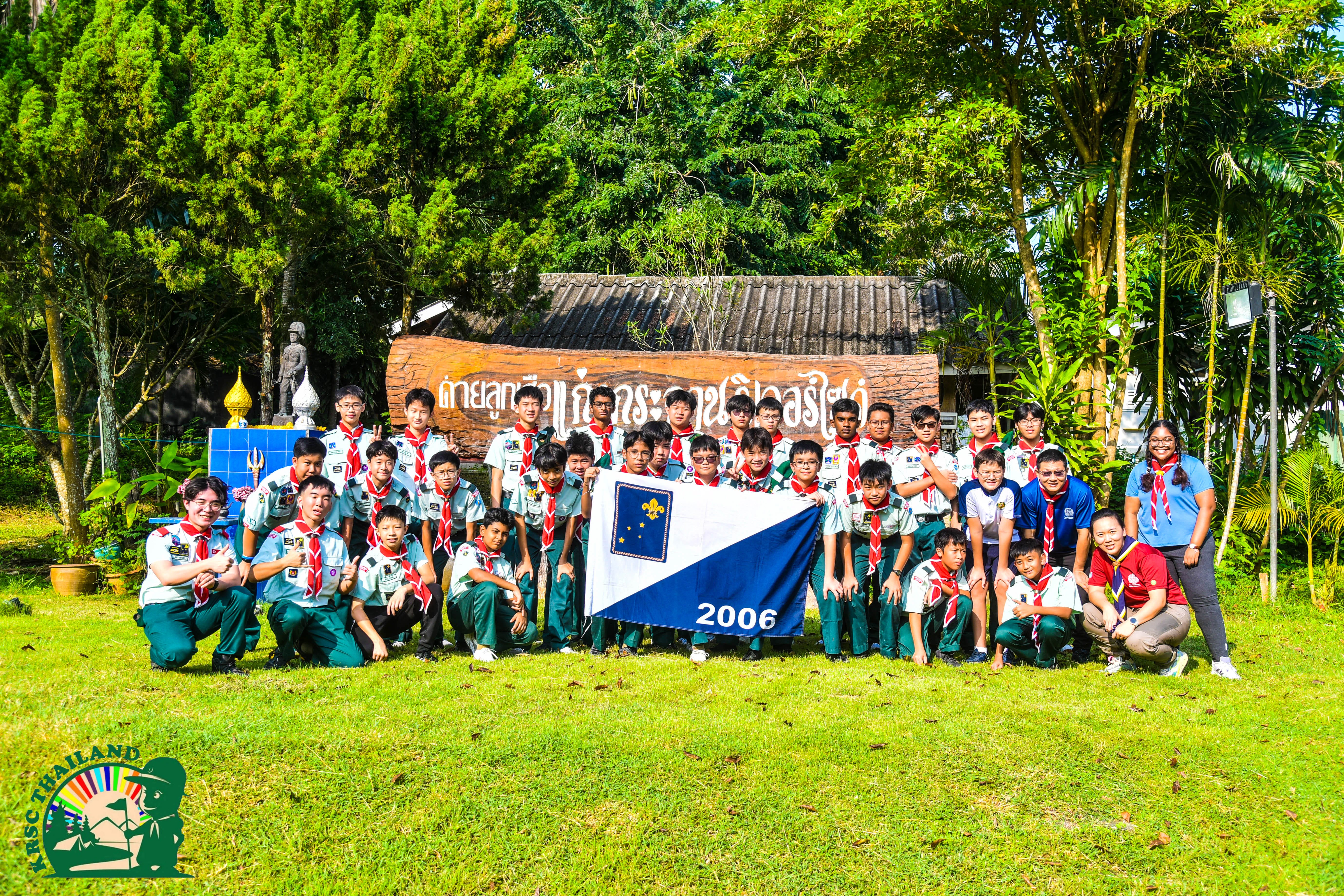 Group in scout uniforms posing on grass with a blue-and-white flag marked 2006, trees and sign behind.