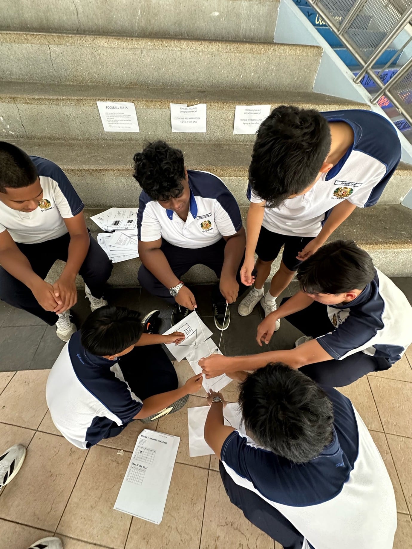 Five students in white and navy uniforms huddle together on steps, reviewing papers and worksheets outdoors.