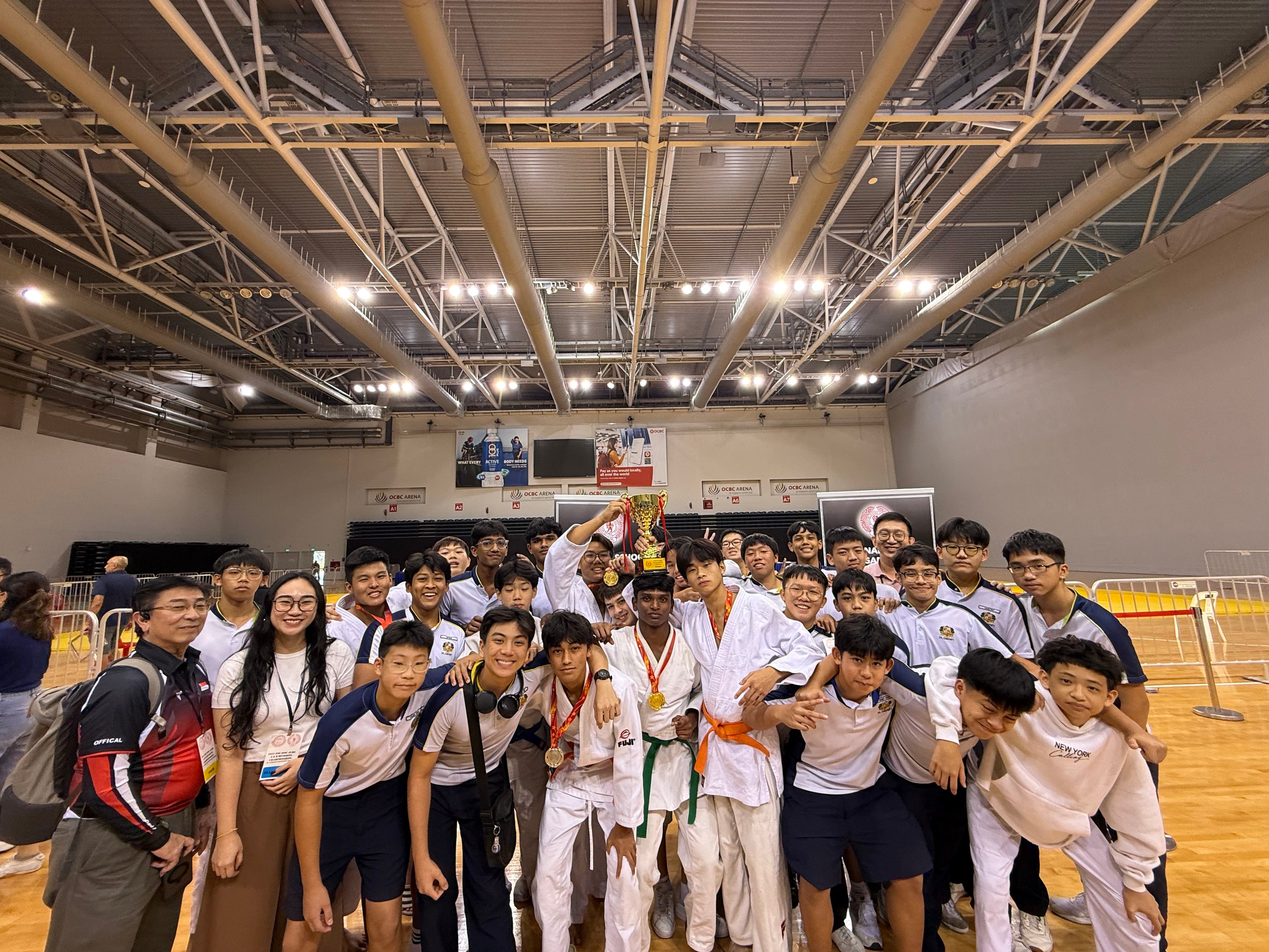 A large group of young male judo athletes and supporters pose with a trophy and medals inside a sports arena.