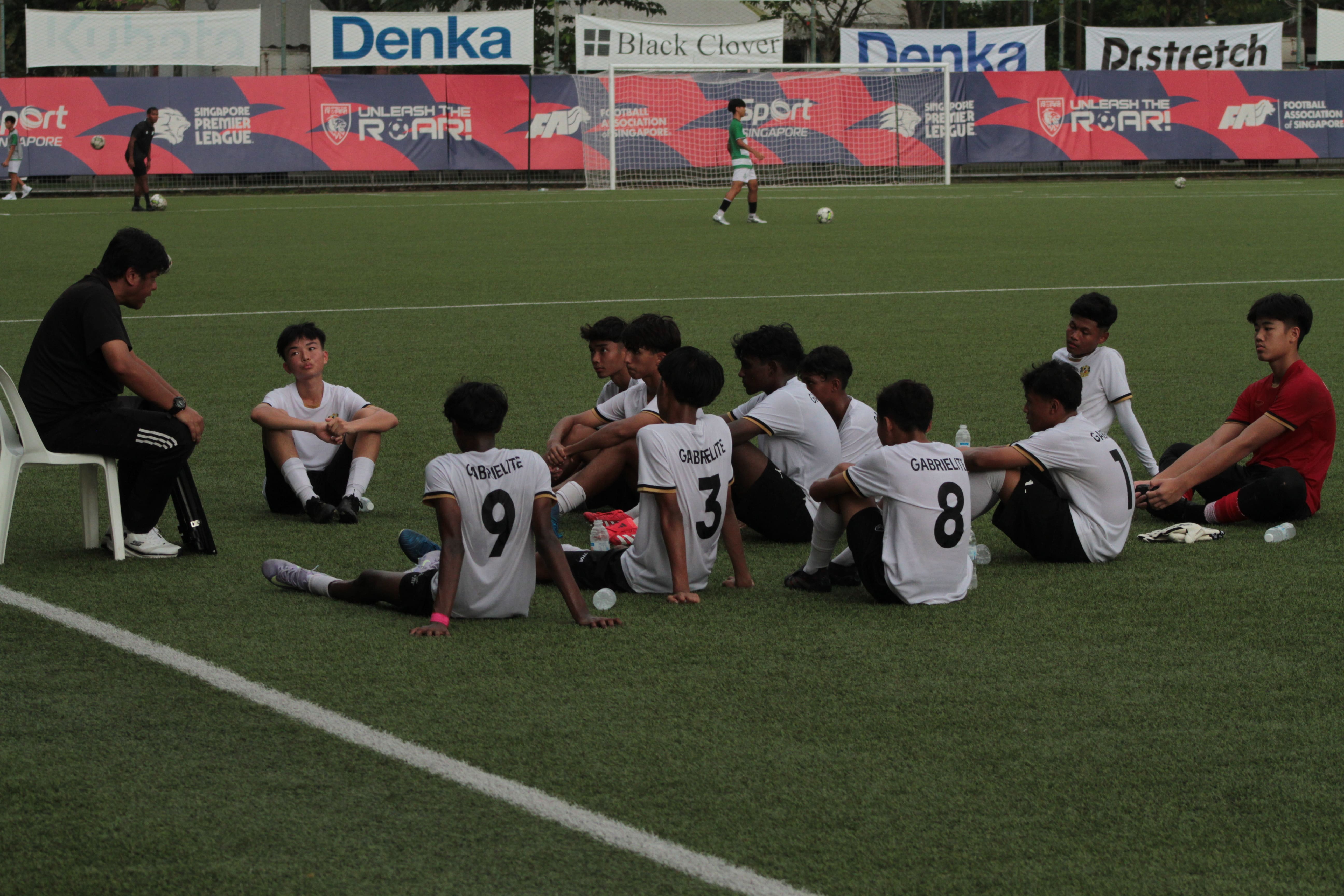 Young footballers in white Gabrielite jerseys sit on a green pitch listening to a coach during a team talk.