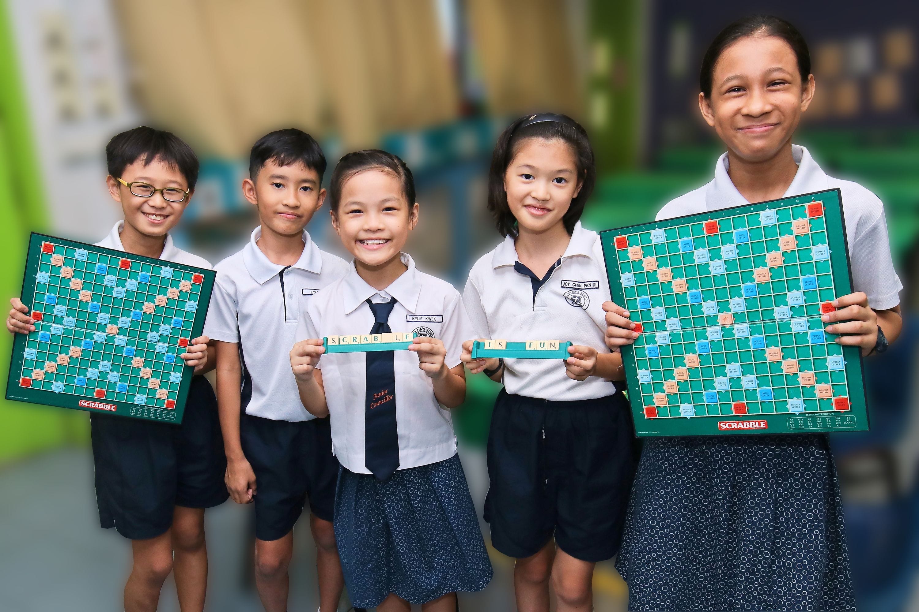 Five smiling school children in uniforms hold Scrabble boards and tiles spelling "Scrabble Is Fun."