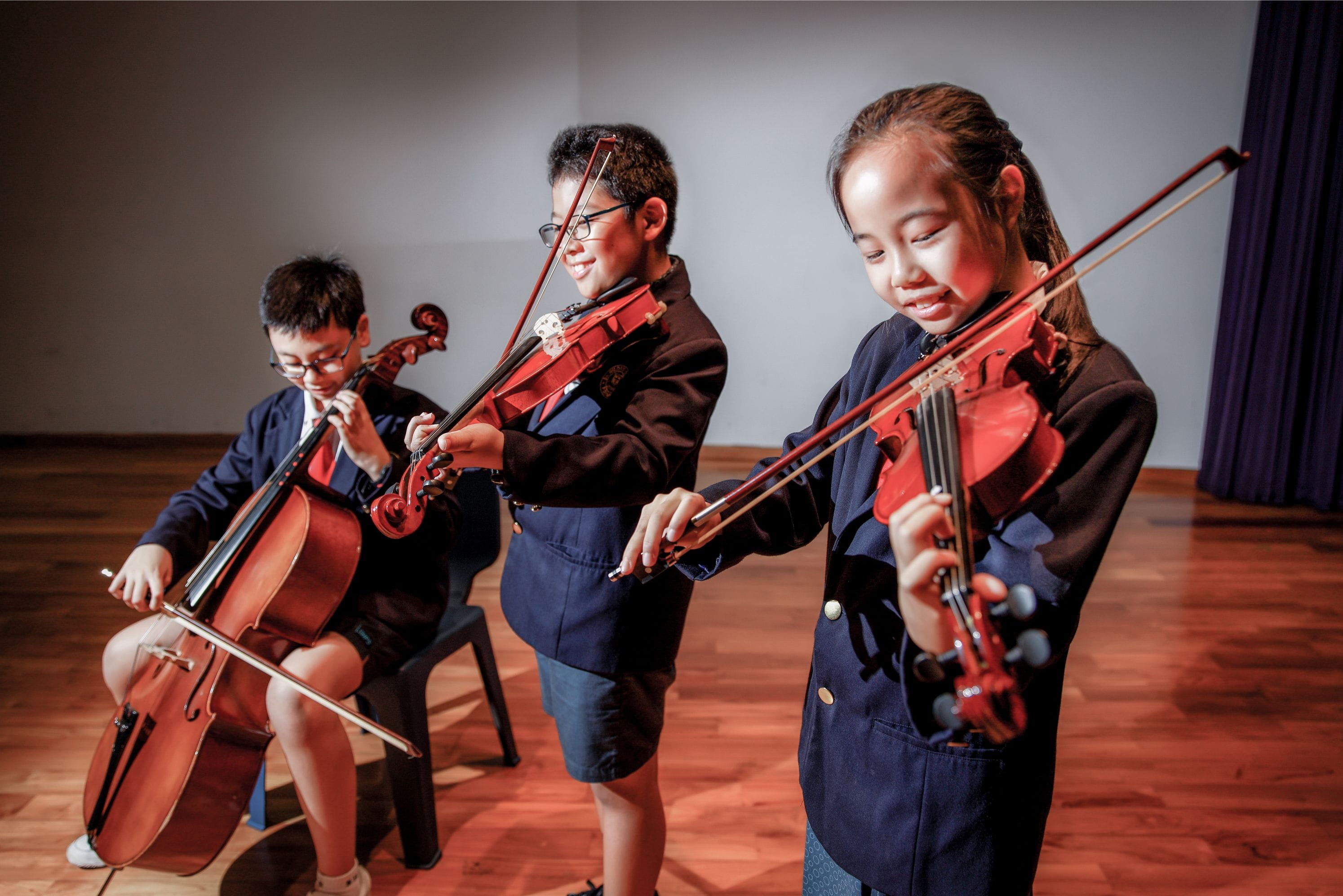 Three children in navy uniforms play string instruments, two with violins and one seated with a cello.