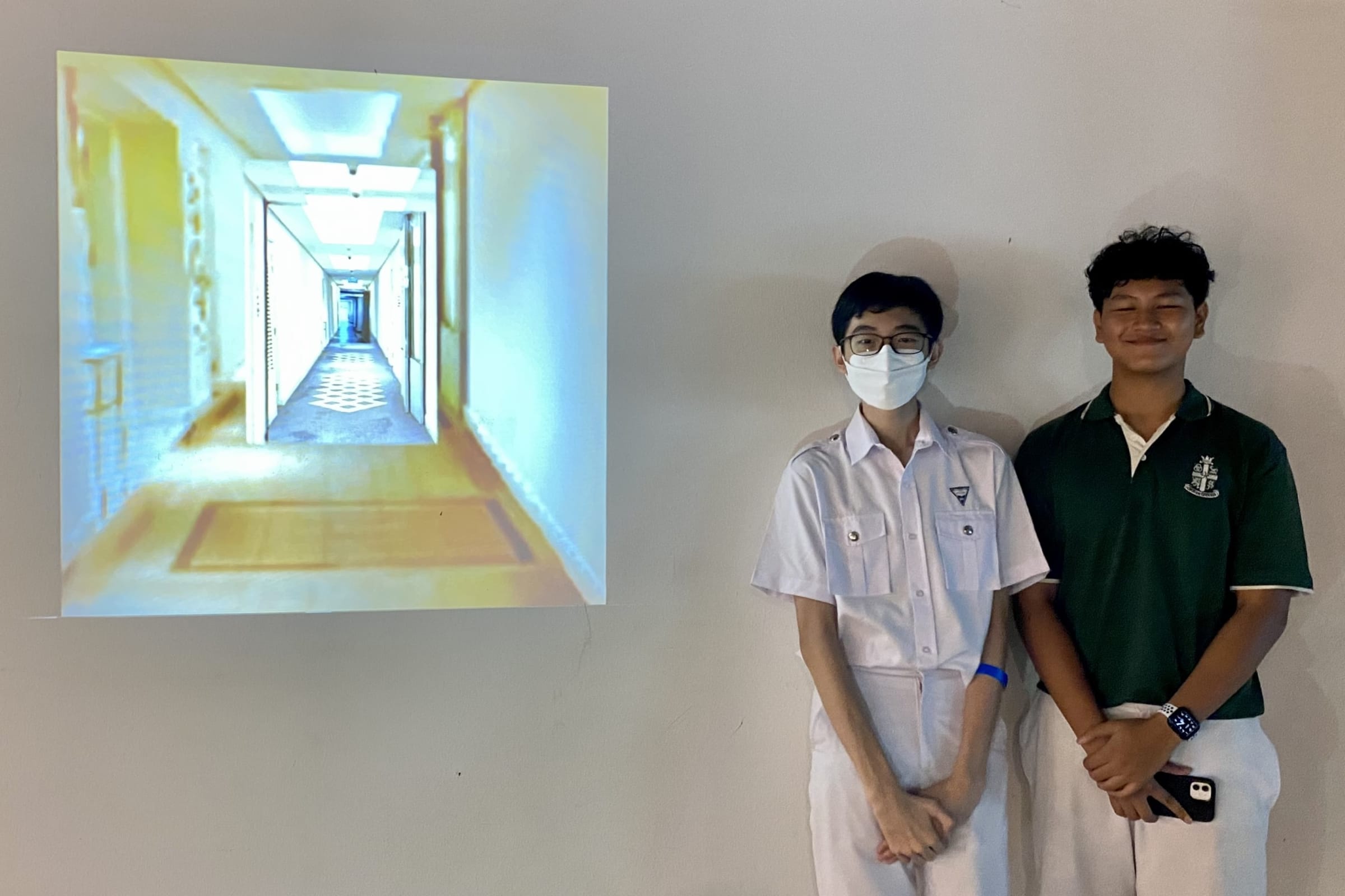Two students in school uniforms stand beside a projected hallway photo on a white wall.