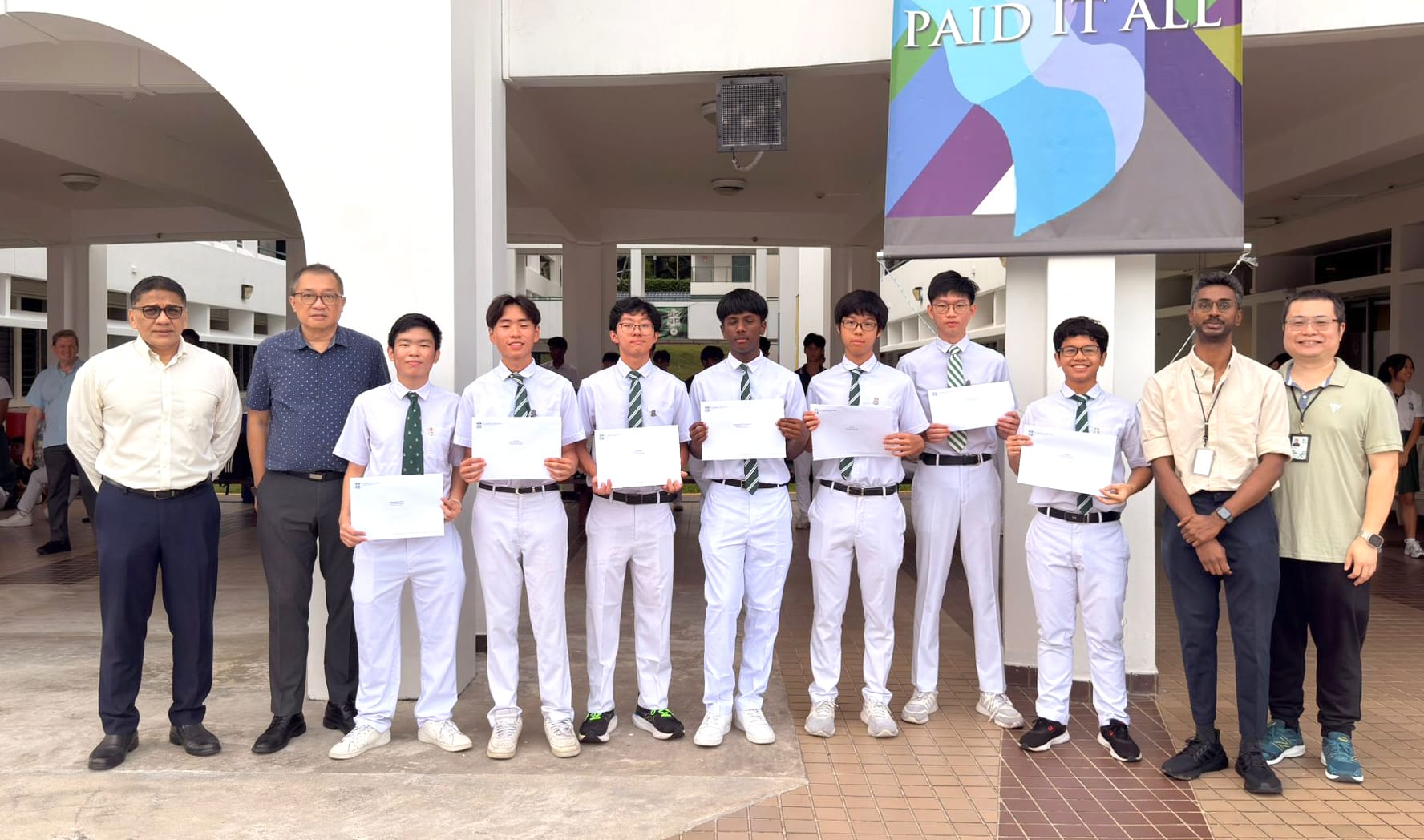 Seven students in white uniforms holding certificates stand with three adults outdoors near a colorful banner.