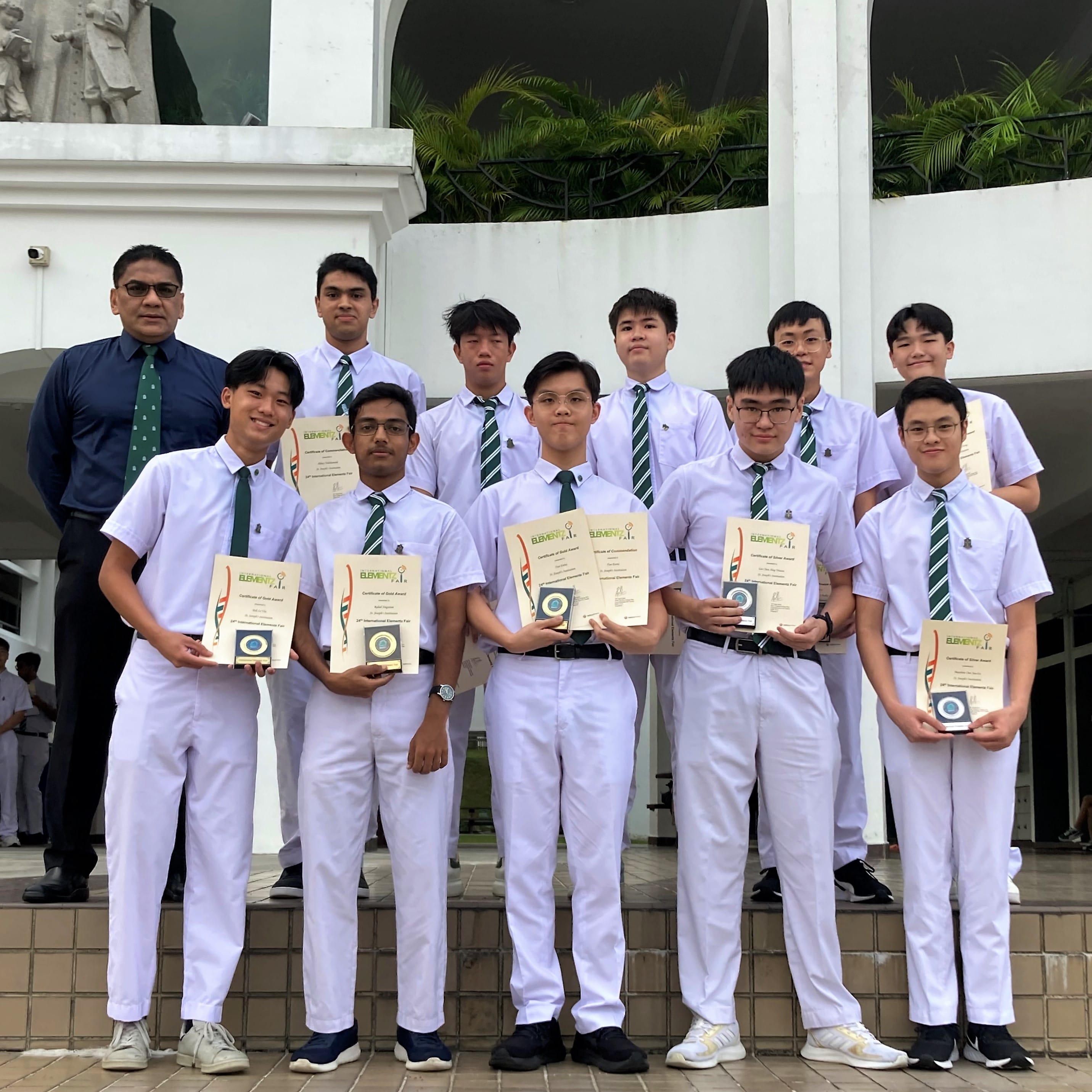 A group of male students in white uniforms with green ties hold award certificates and medals, posing with a teacher outdoors.
