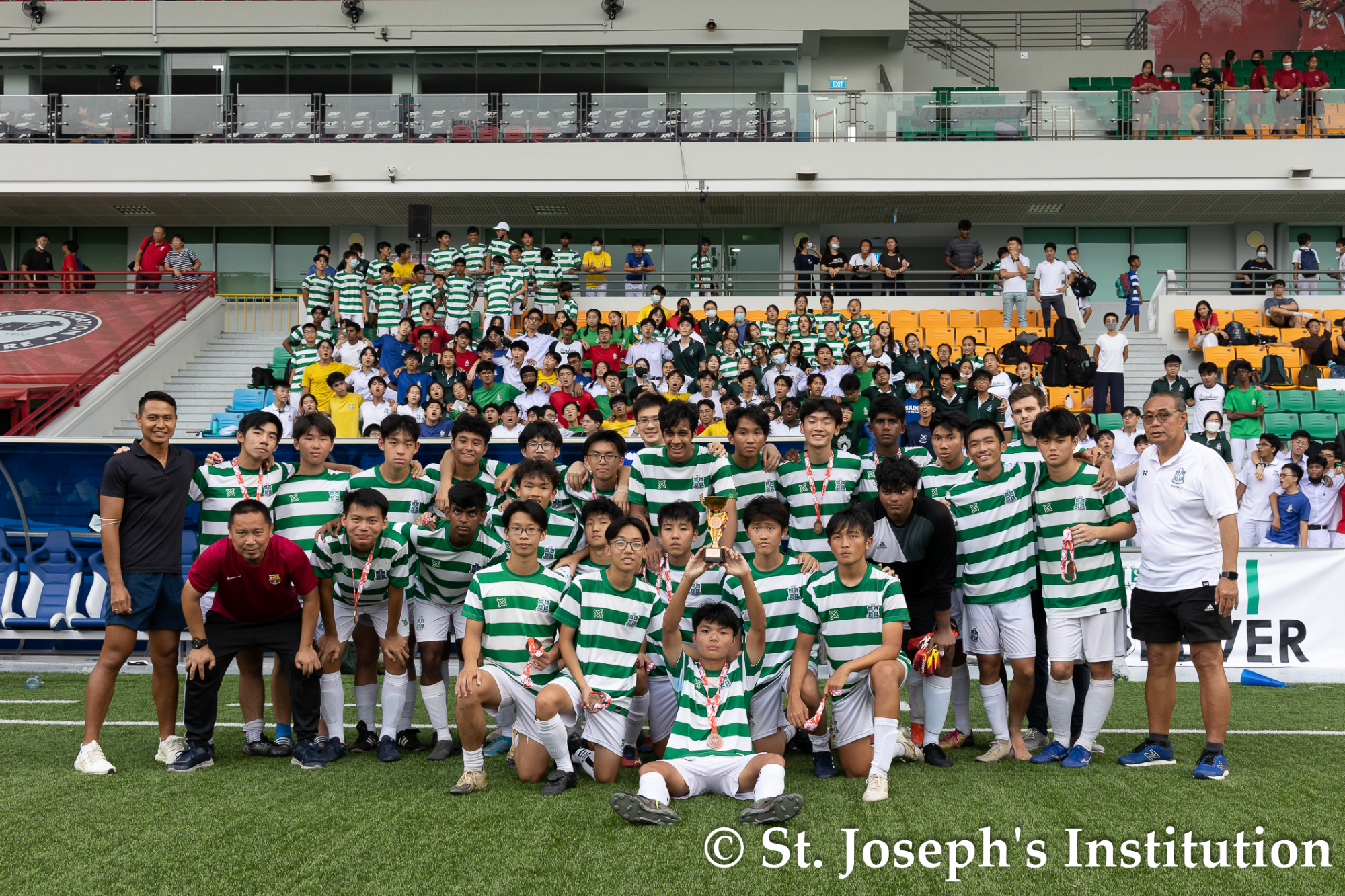 Soccer team in green-and-white striped jerseys pose with a trophy on a field, crowd in stadium seats behind.