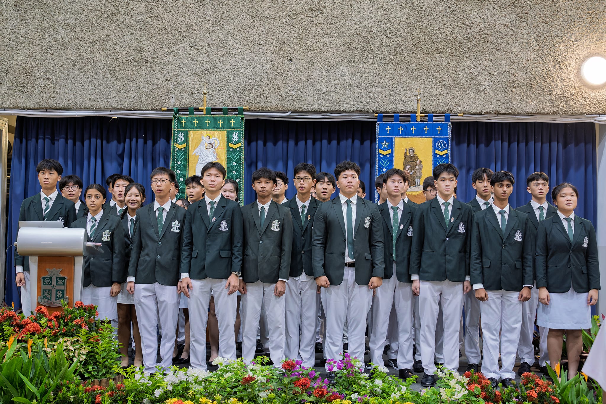 Students in green blazers and white pants stand on a stage with religious banners and flowers in front.