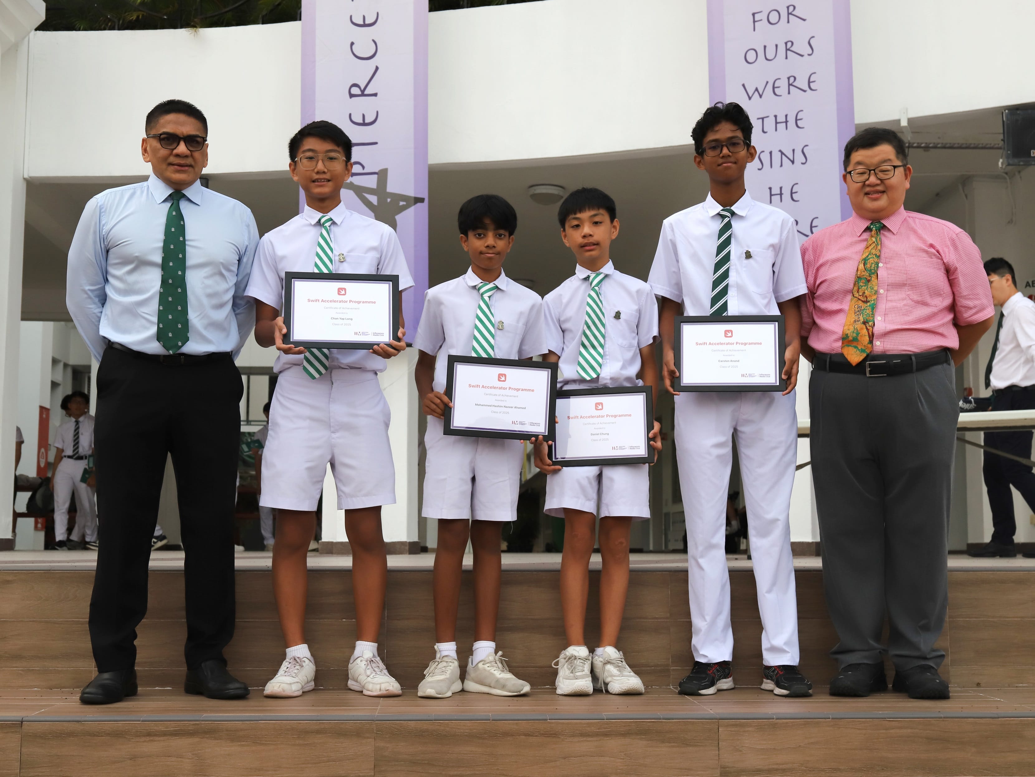 Four uniformed school boys holding framed certificates stand on a stage flanked by two adult men in shirts and ties.