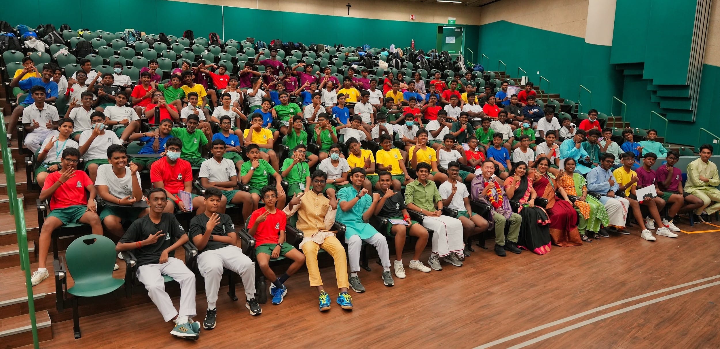 Large group of students and adults seated in tiered auditorium seating, wearing colorful shirts, posing for a photo.