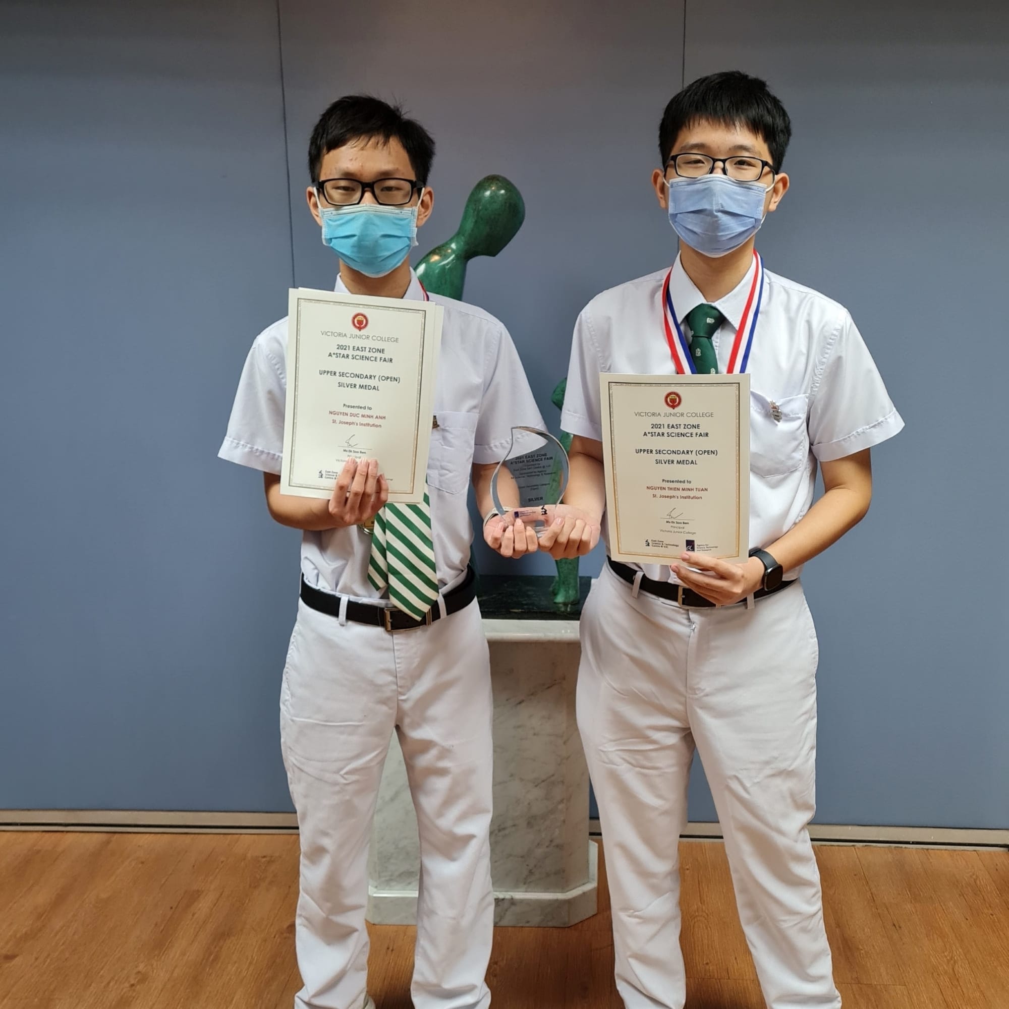 Two students hold a glass trophy and their 2021 East Zone A*STAR Science Fair Upper Secondary Silver Medal certificates.
