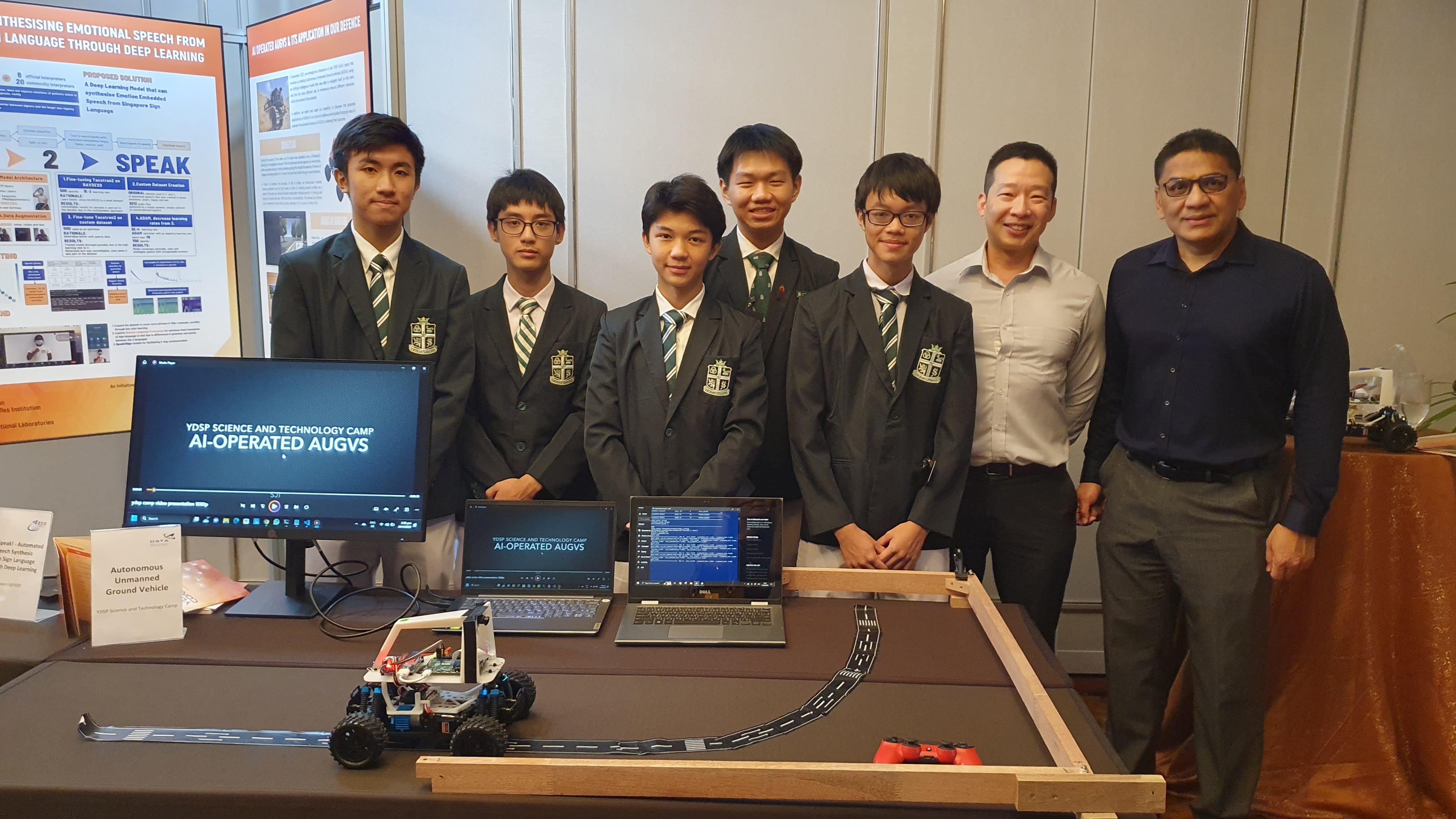 Seven people stand behind a table displaying a small robot vehicle, laptops, and a monitor showing AI-Operated AUGVS.