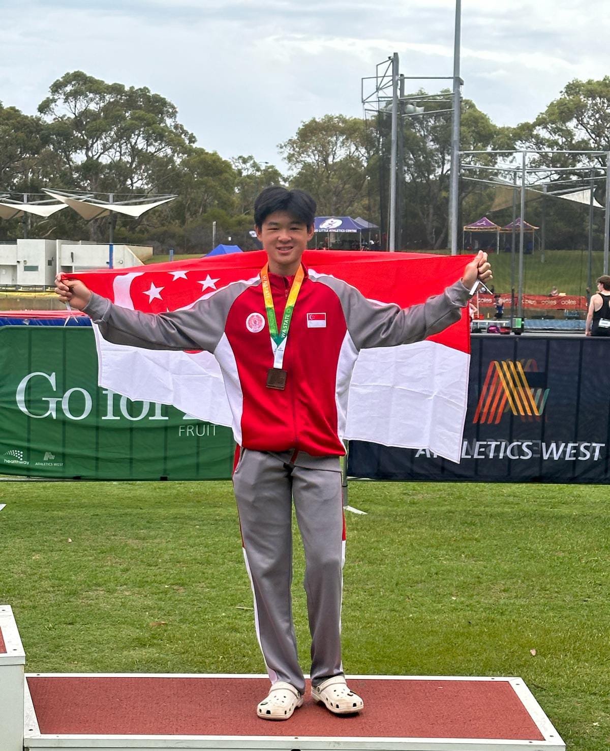 Young man wearing a medal stands on a podium holding the Singapore flag wide at an Athletics West event.