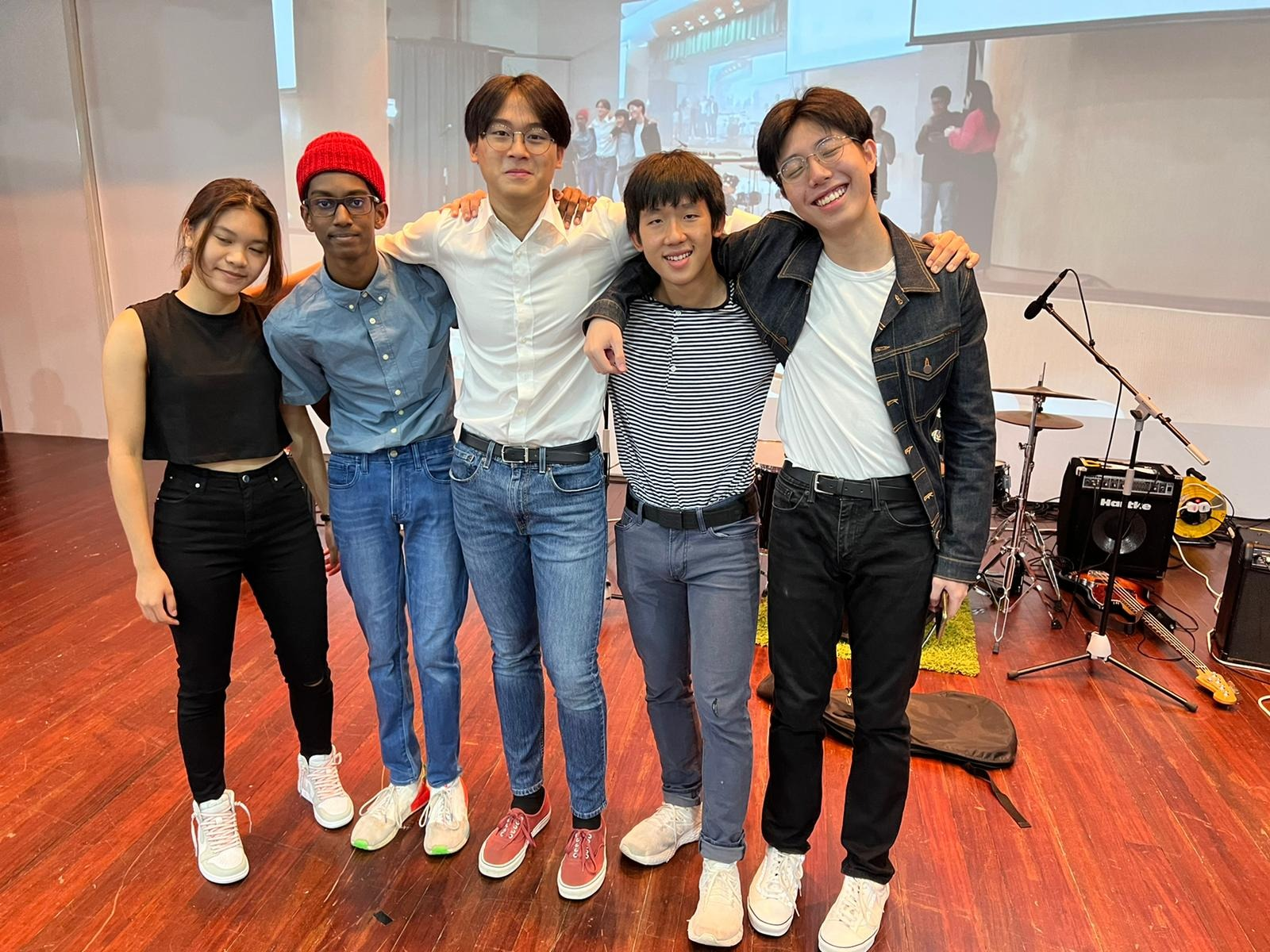 Five young people pose together smiling on a wooden stage with drums and an amp visible nearby.