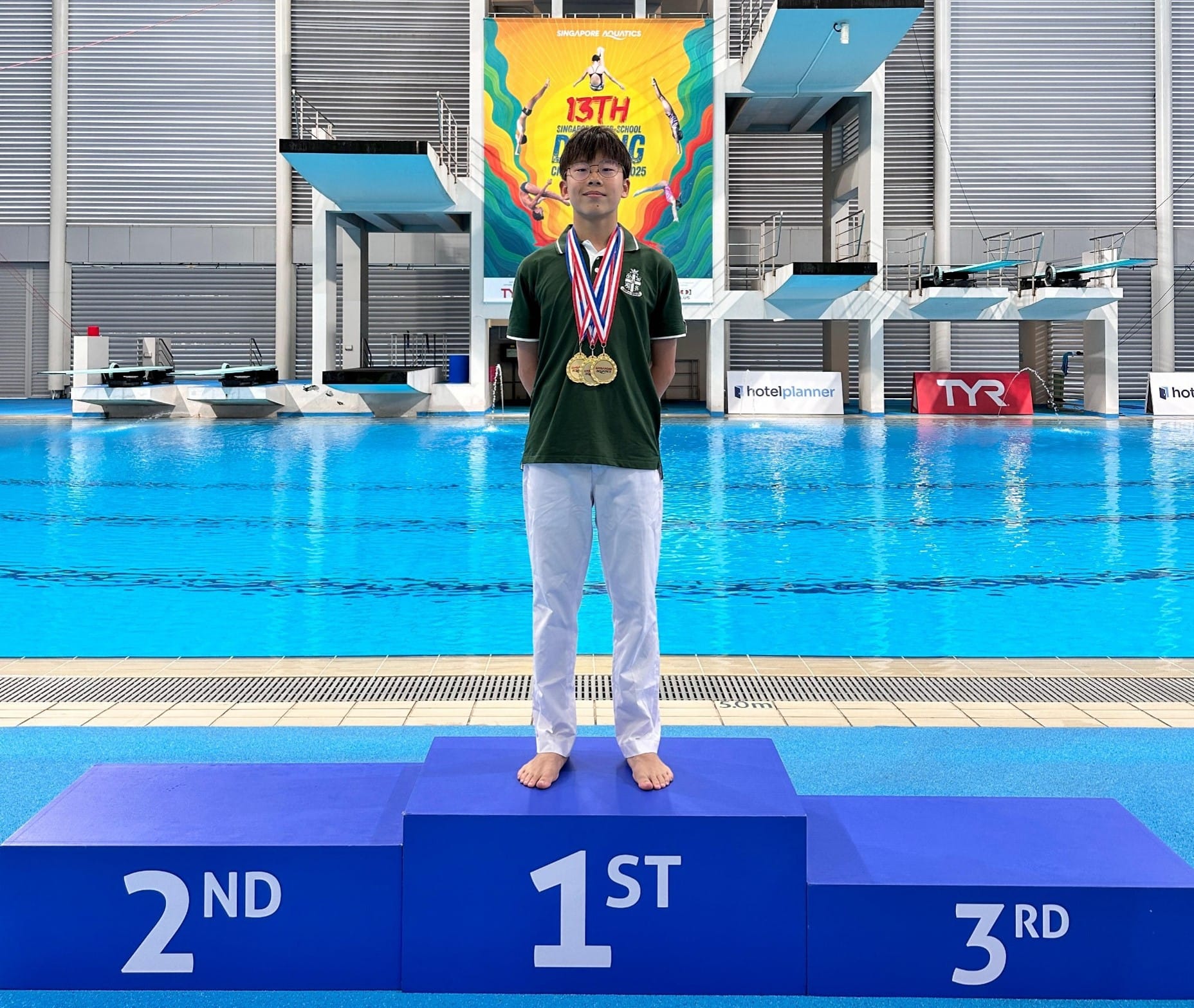 A young boy wearing gold medals stands on the 1st place podium at the 13th Singapore School Diving Championship.