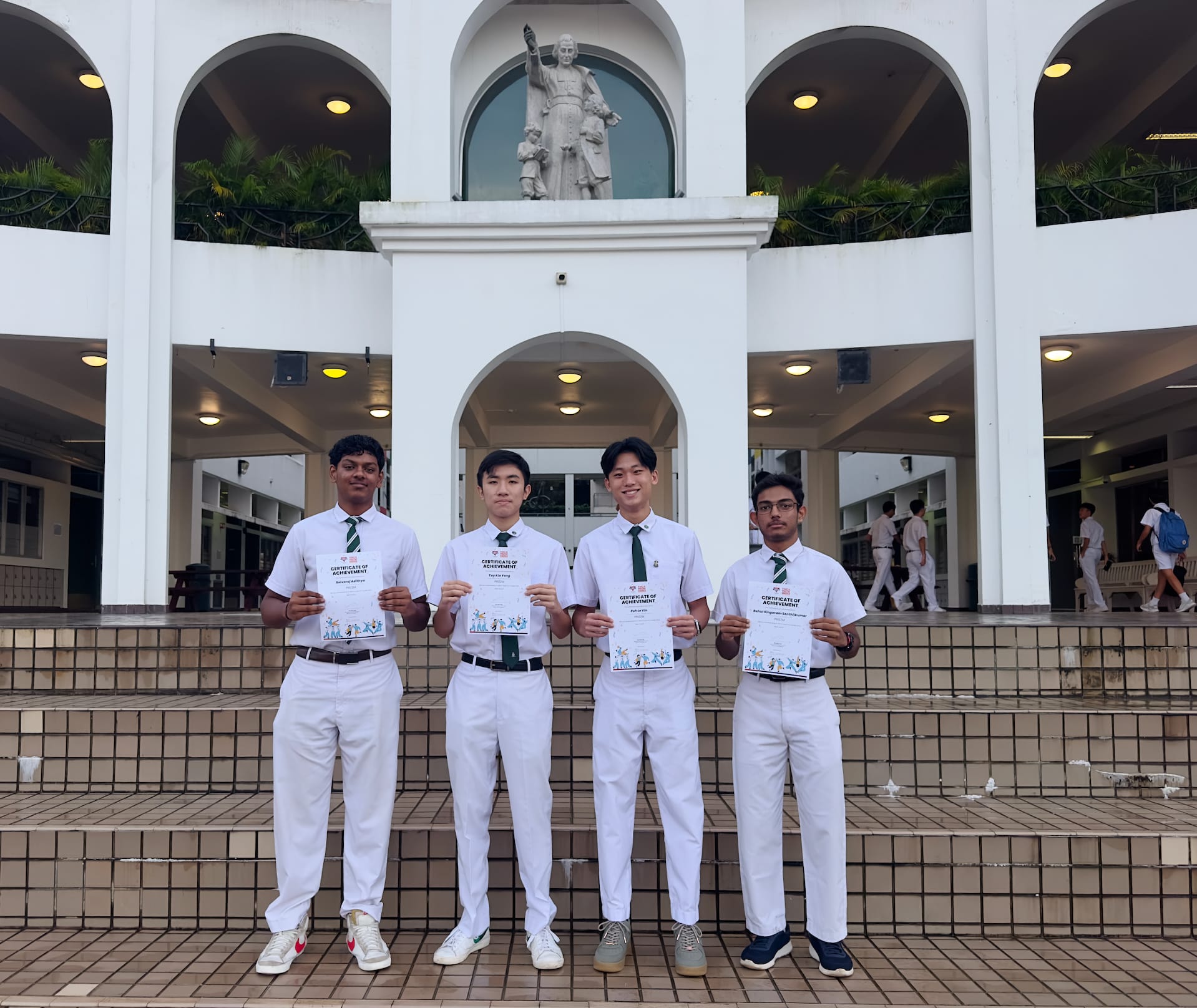Four male students in white uniforms hold certificates of achievement in front of a building with arches and a statue.