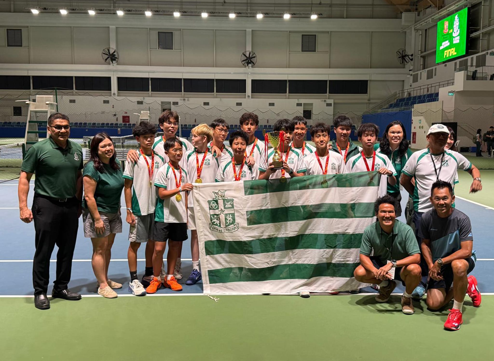 A youth tennis team in green and white uniforms poses with medals, a trophy, and a striped school flag on an indoor court.