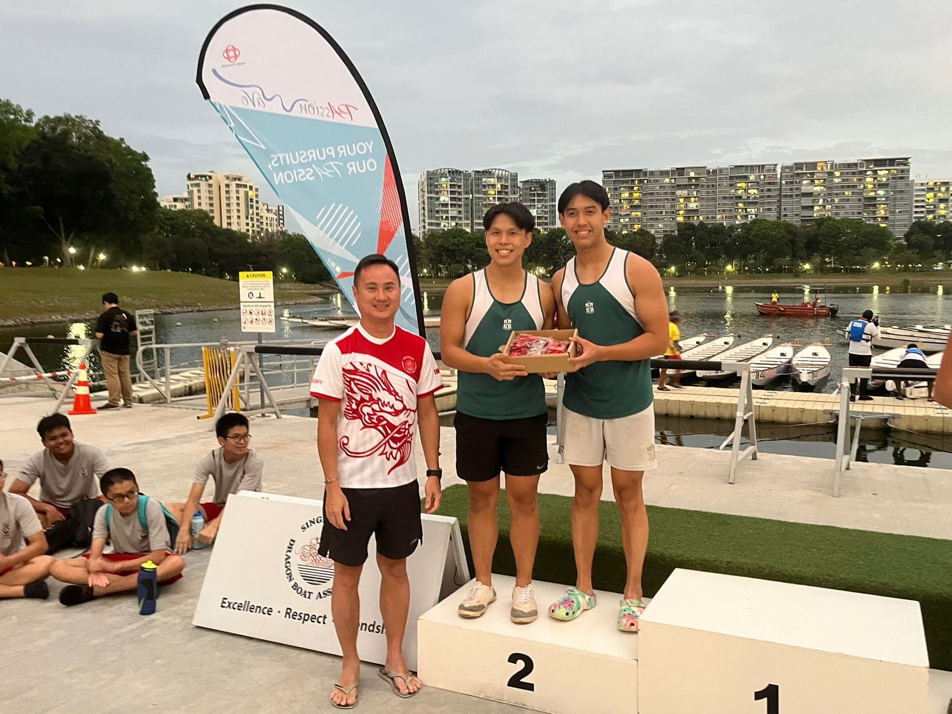 Three people stand on a podium at a Singapore Dragon Boat Association event, holding a box near a waterway.
