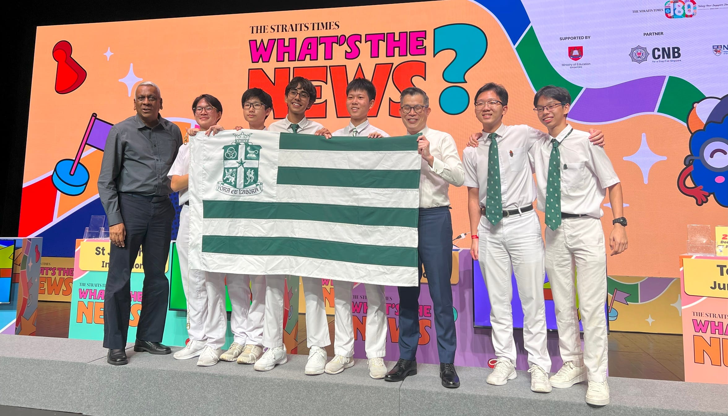 A group of students and adults pose on stage holding a green and white striped school flag at The Straits Times What's The News event.