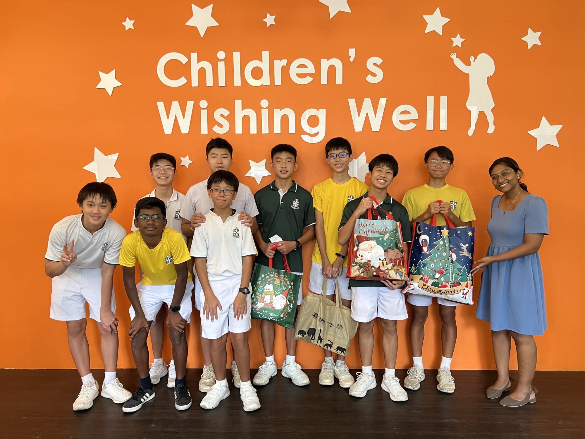 Ten students pose in front of an orange Children's Wishing Well wall, holding Christmas gift bags.