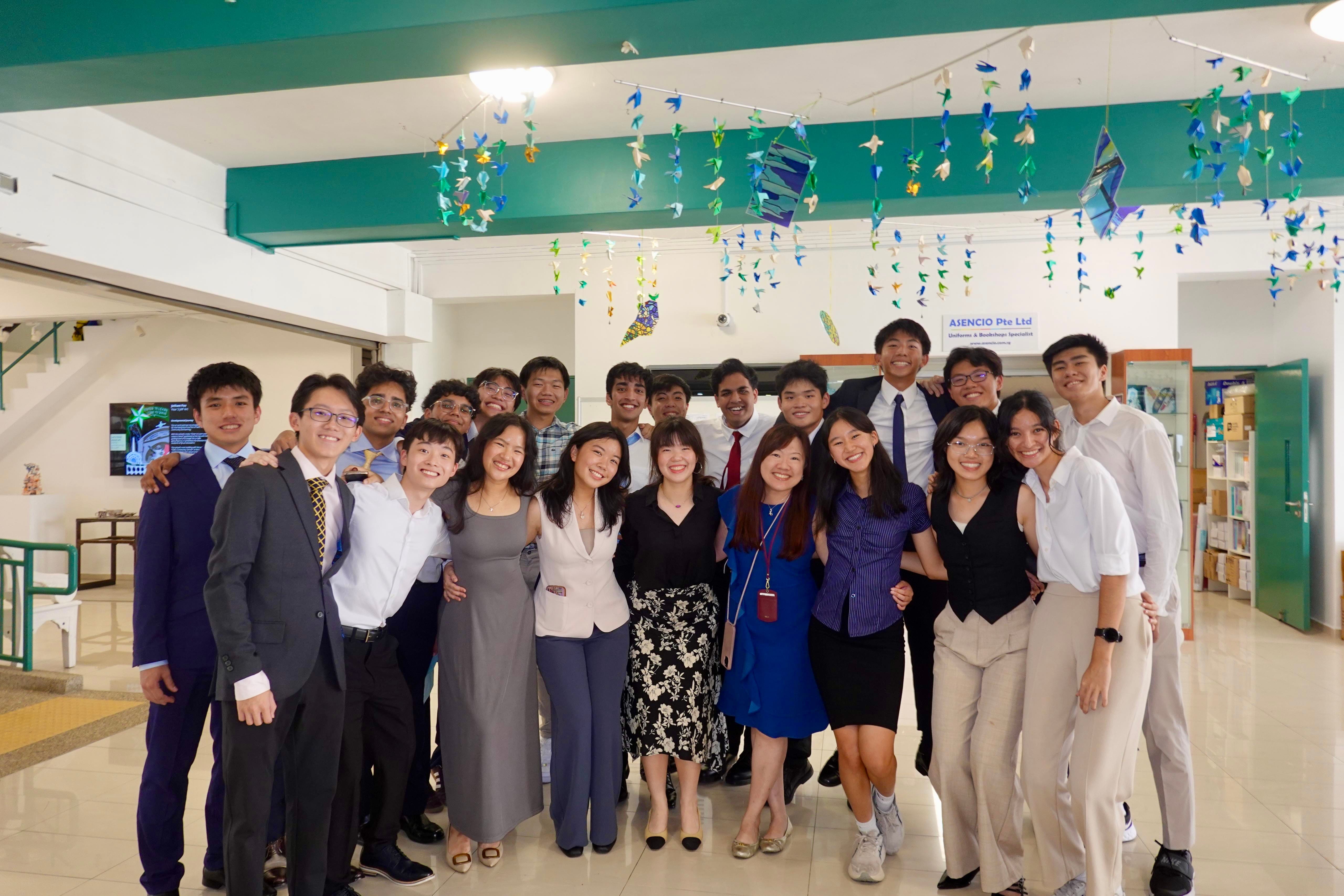 A large group of formally dressed young people smiling together indoors under hanging origami paper crane decorations.