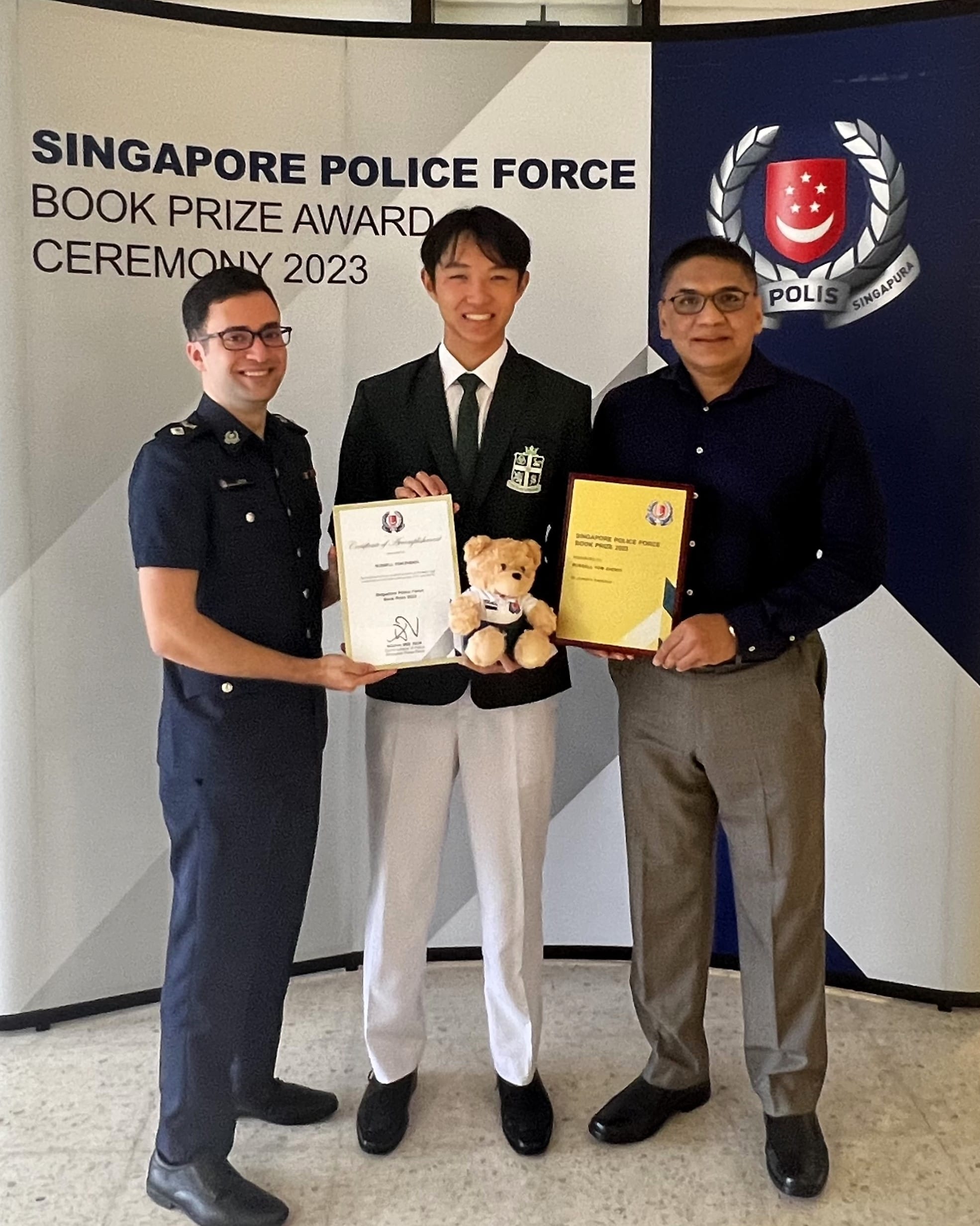 Three men pose at the Singapore Police Force Book Prize Award Ceremony 2023, holding a certificate, plaque, and teddy bear.