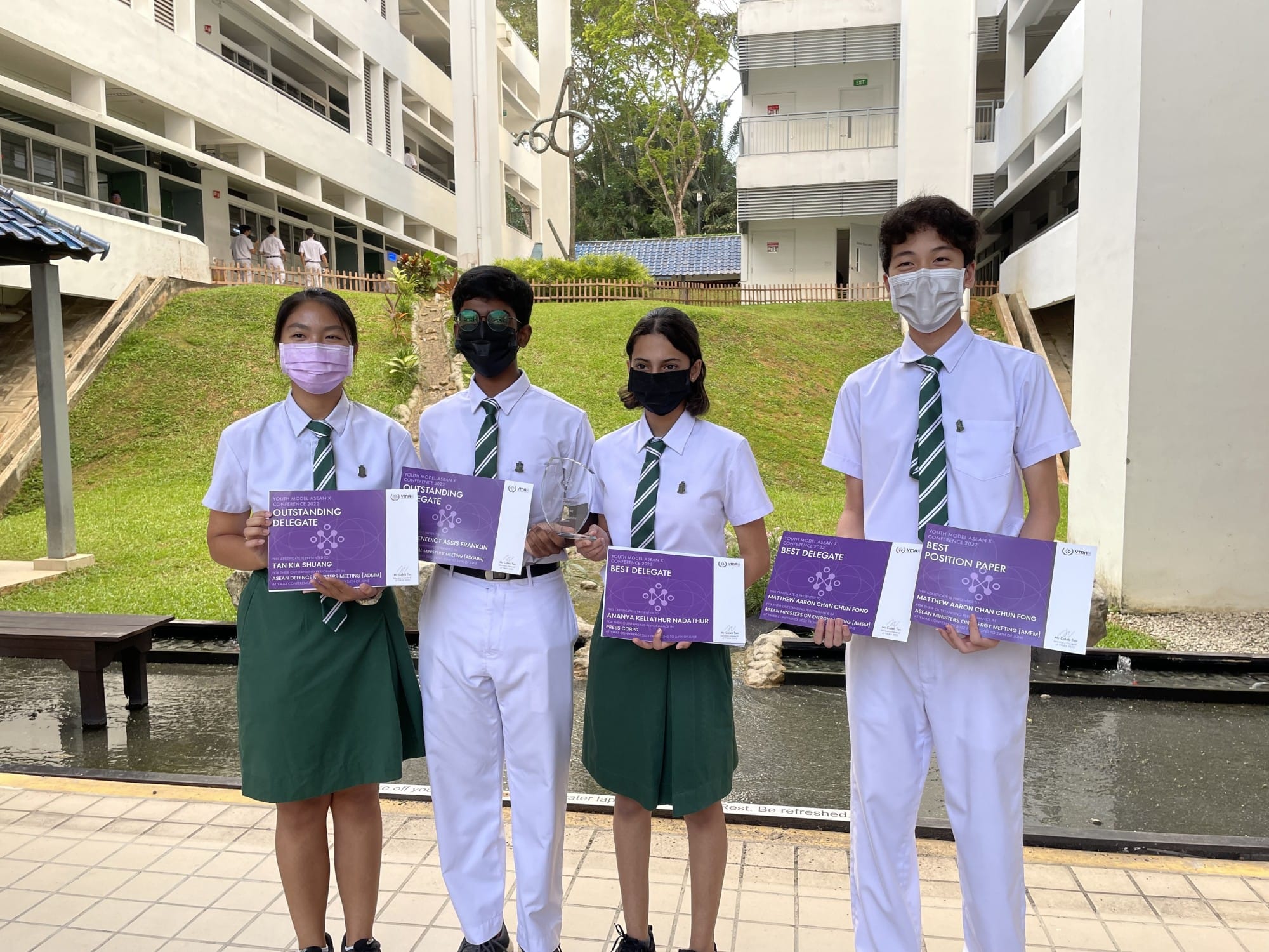 Four masked students in white uniforms hold purple award certificates outdoors at a school campus.