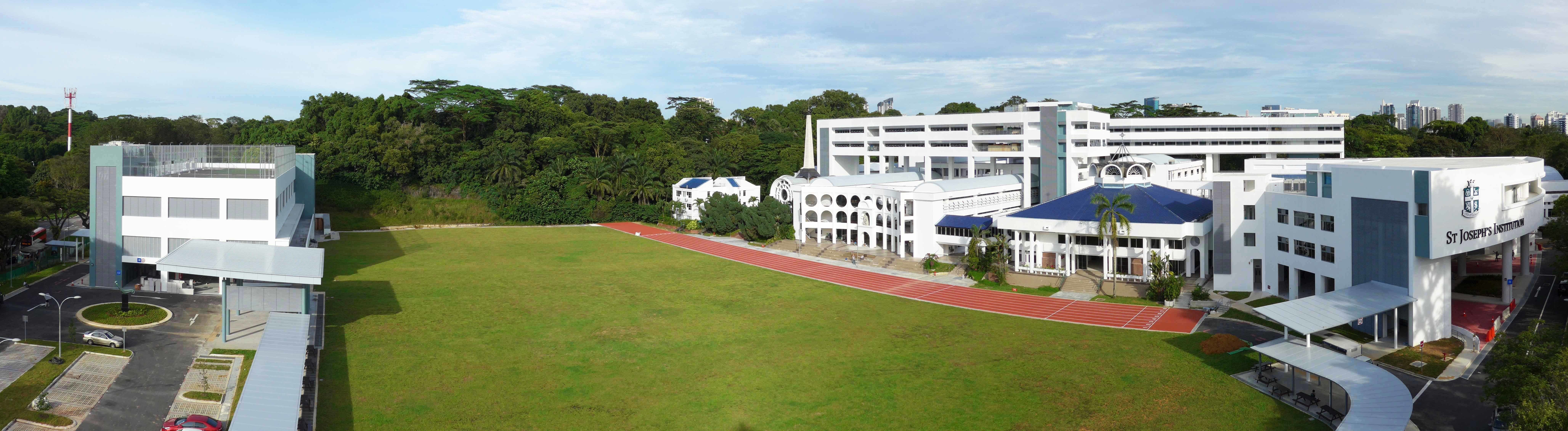 Wide aerial view of school campus with large grass field, red running track, and white buildings.