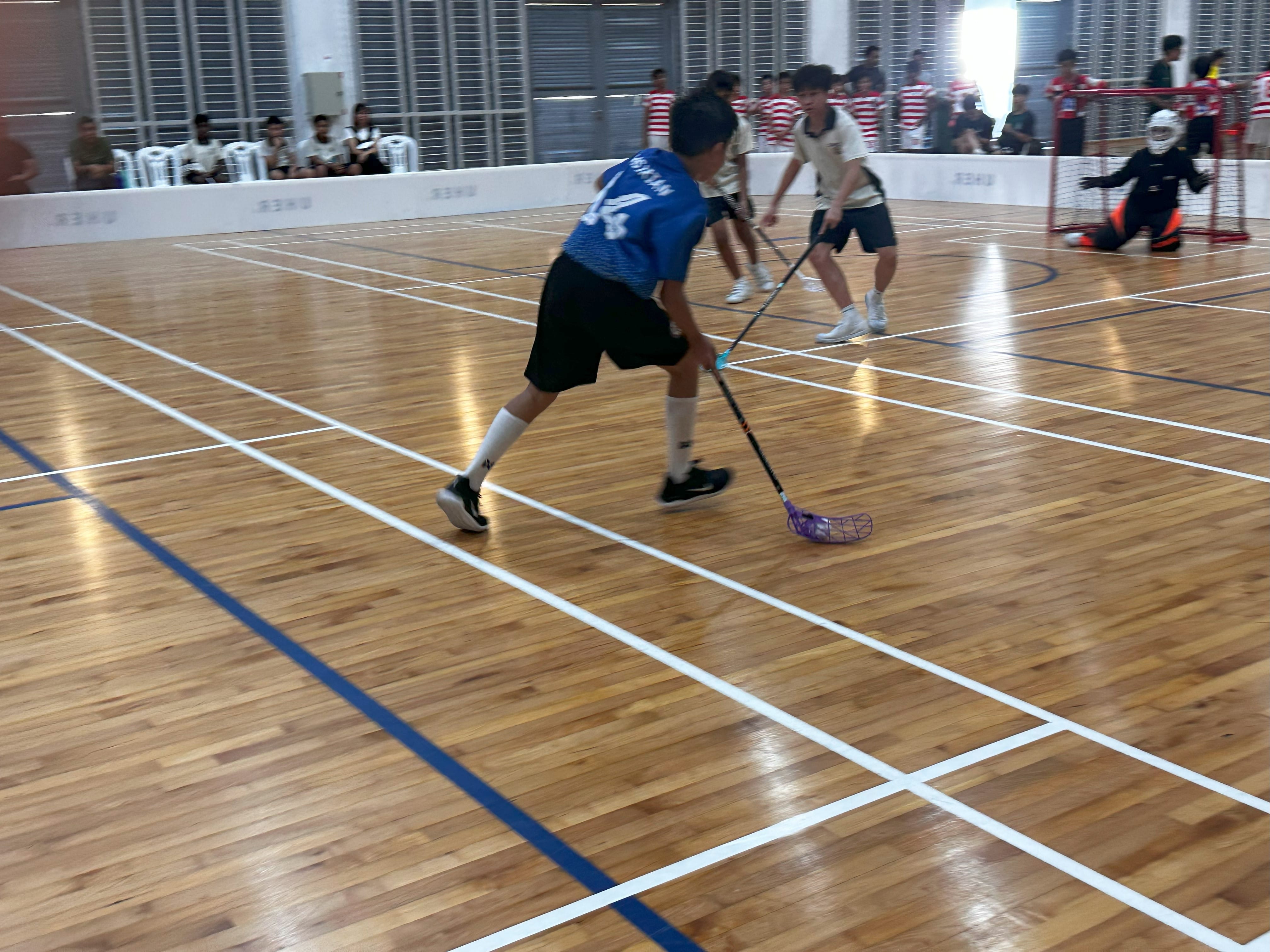 Youth players compete in an indoor floorball match on a wooden court, with a goalkeeper near the red goal.