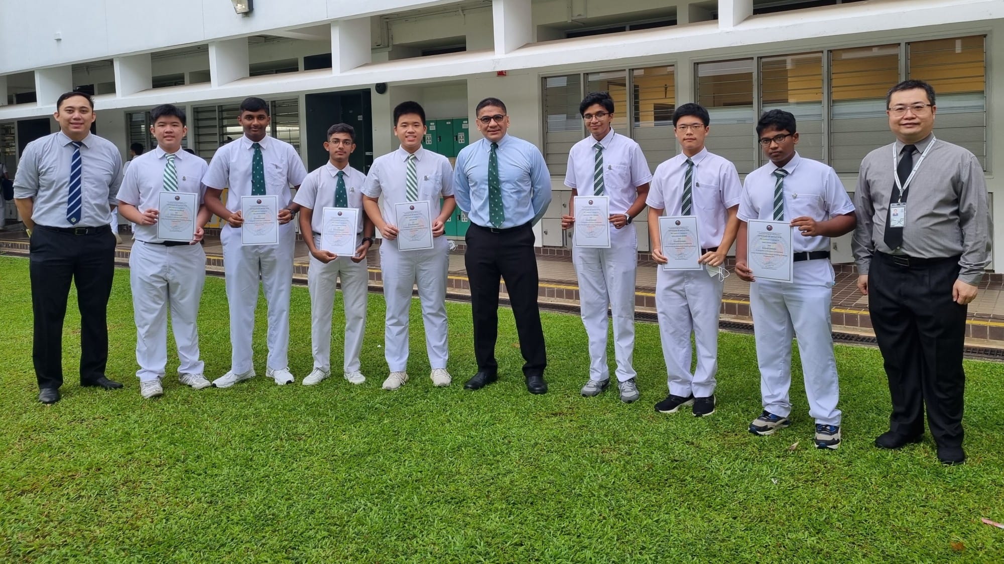 Ten people in white uniforms and ties stand on grass outside a school building, with seven holding certificates.