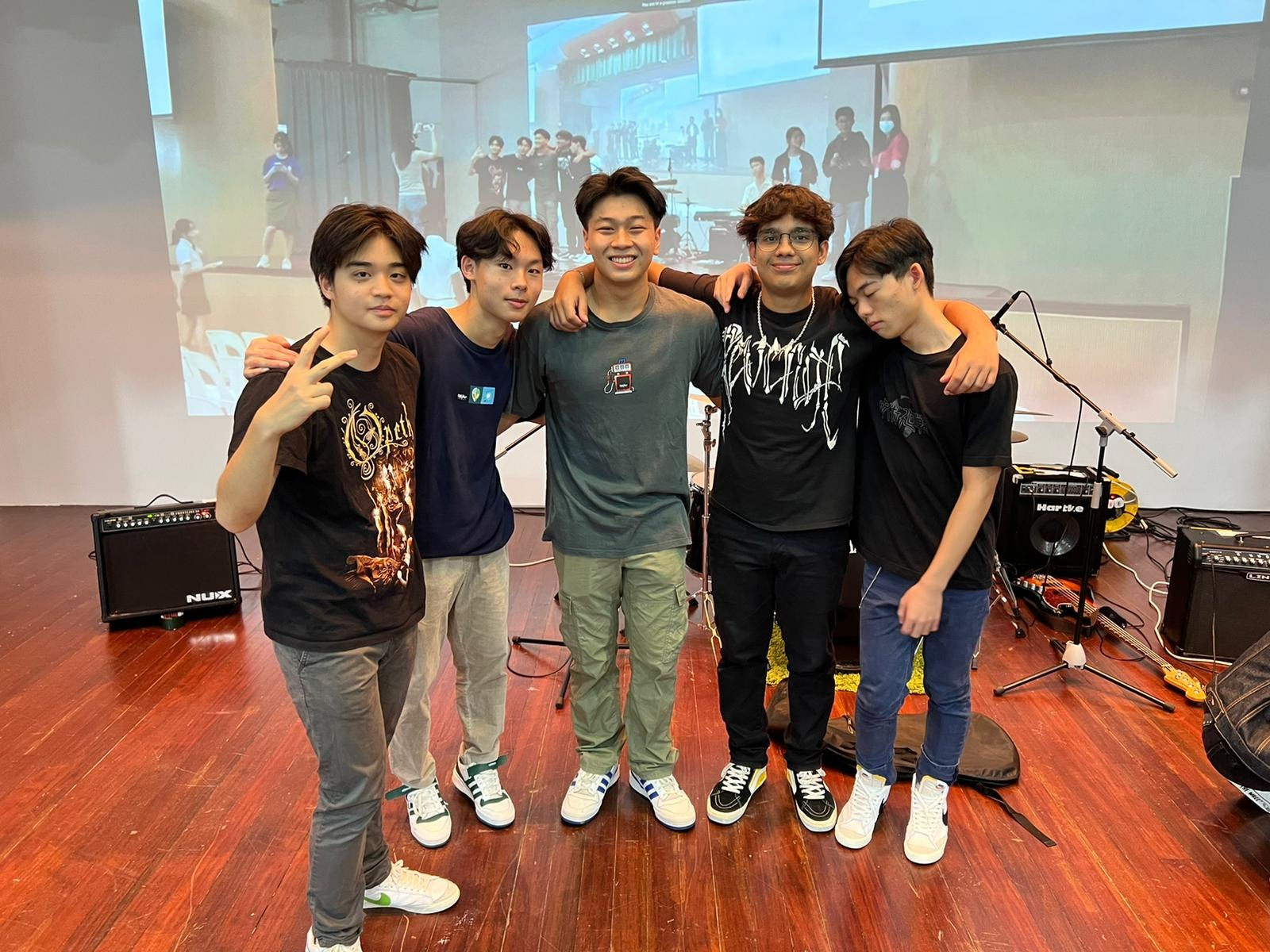 Five young men pose together on a stage with music equipment and a projection screen behind them.
