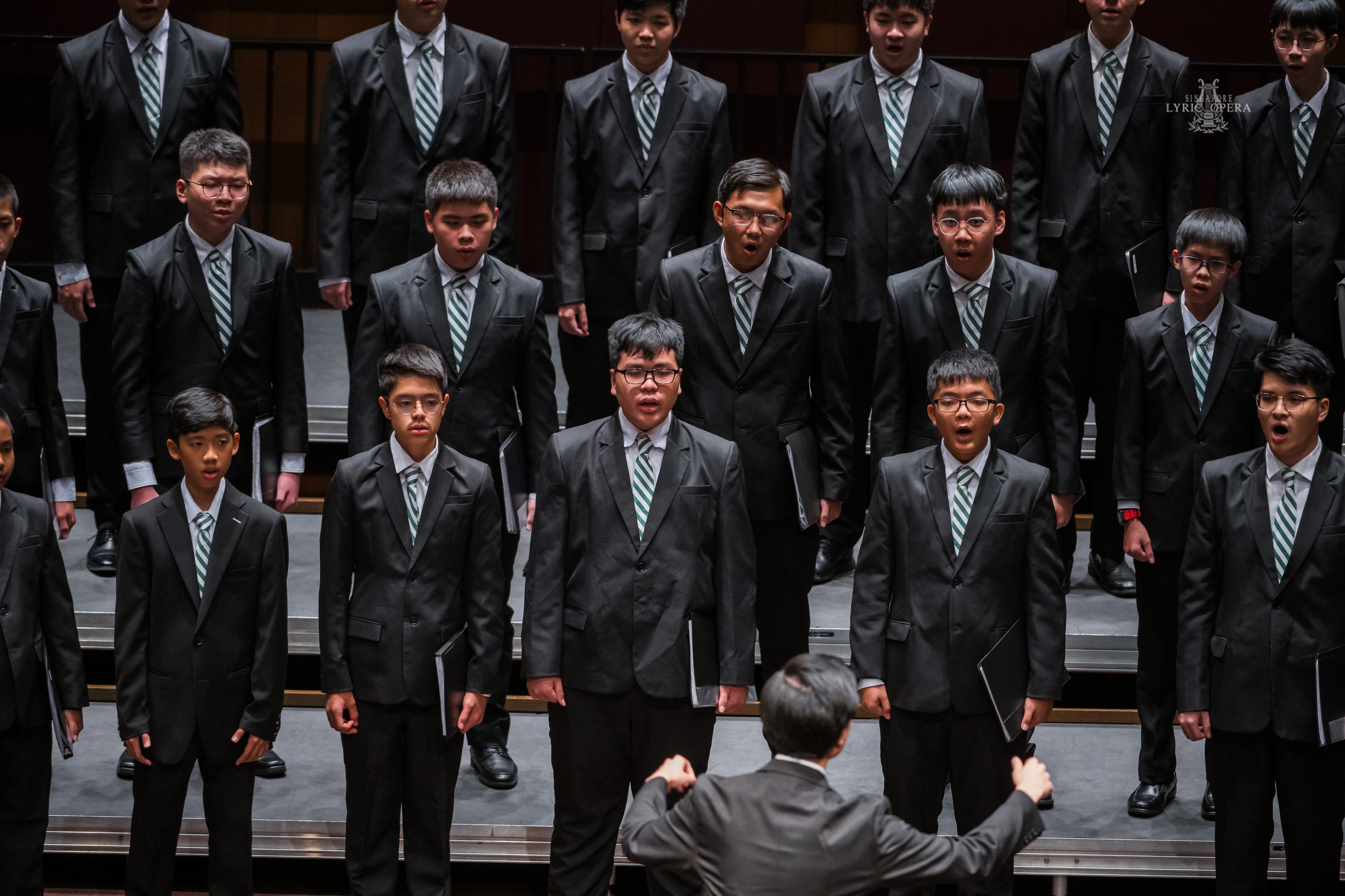 A conductor leads a boys choir in dark suits with green striped ties, singing on tiered risers.