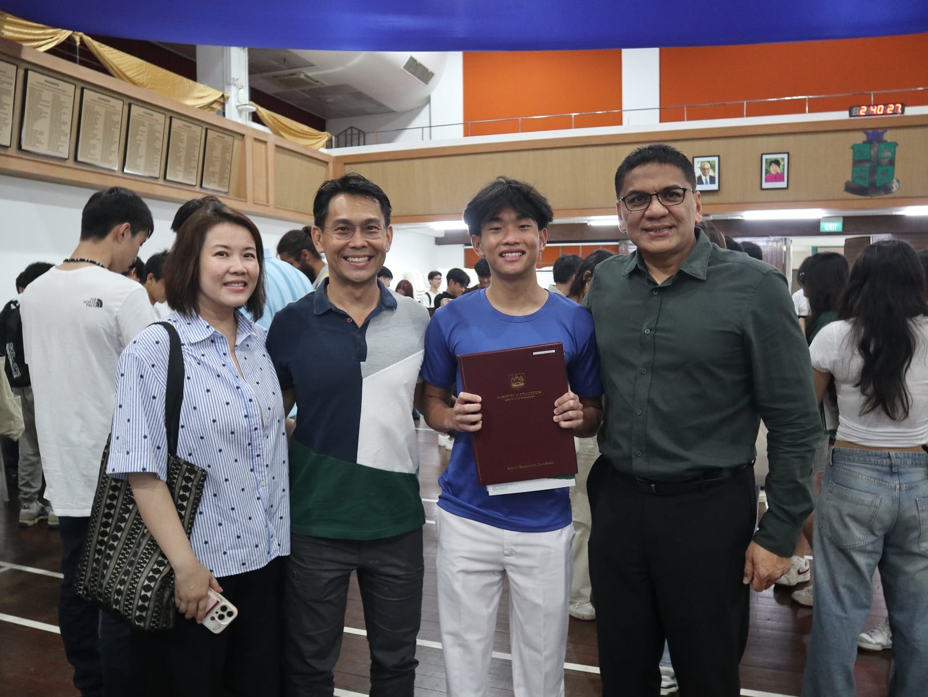 Four smiling people pose together indoors; a young man holds a red certificate folder at what appears to be a school event.