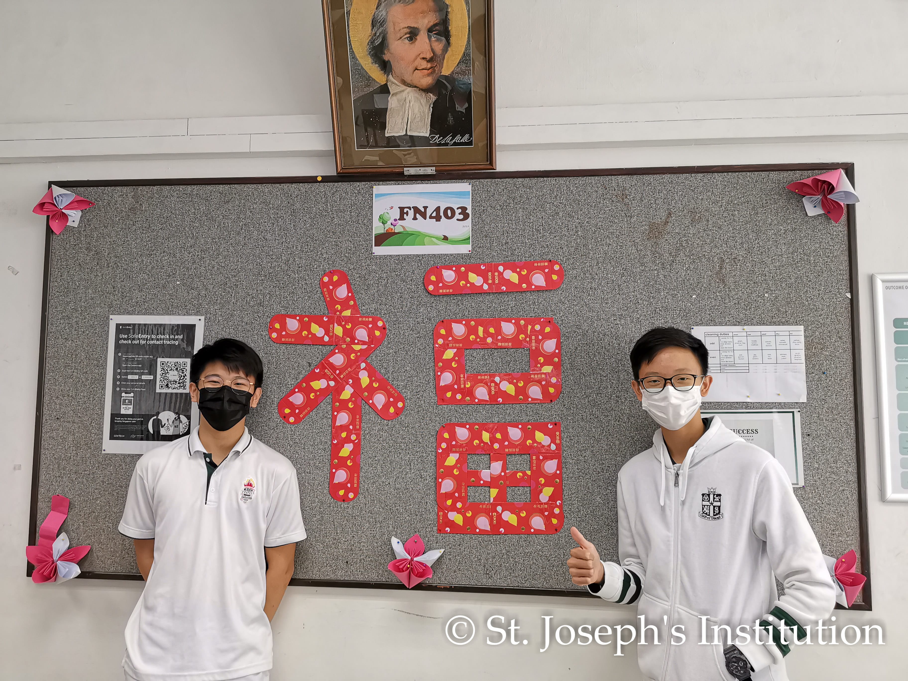Two masked students in white uniforms stand before a board with a large red Chinese character made from red packets.