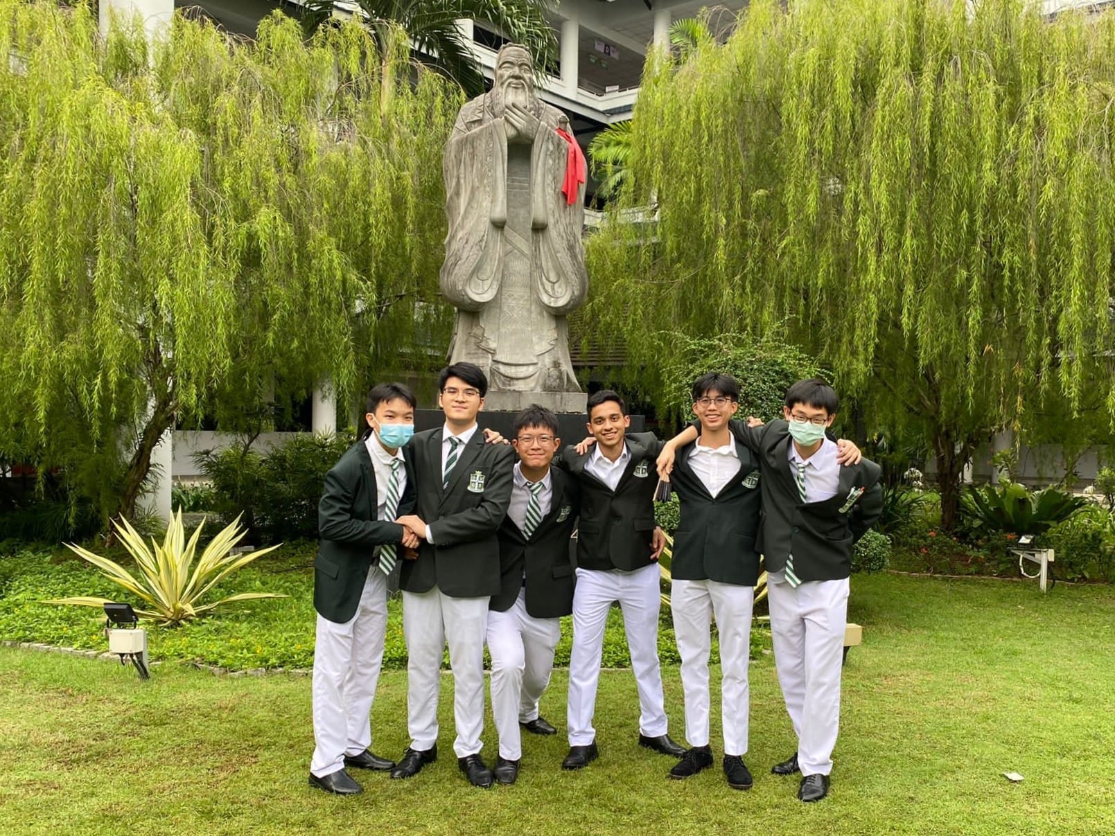 Six male students in green blazers and white pants pose on grass before a large stone robed figure statue.