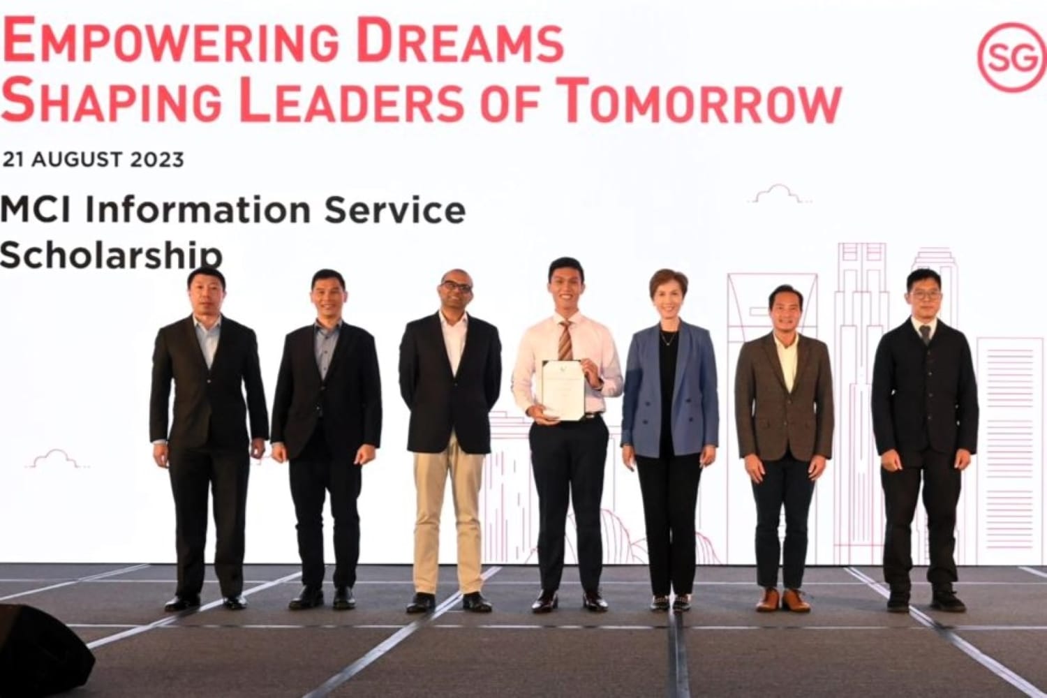 Seven people on stage for the MCI Information Service Scholarship, 21 August 2023, with the recipient holding a certificate at center.