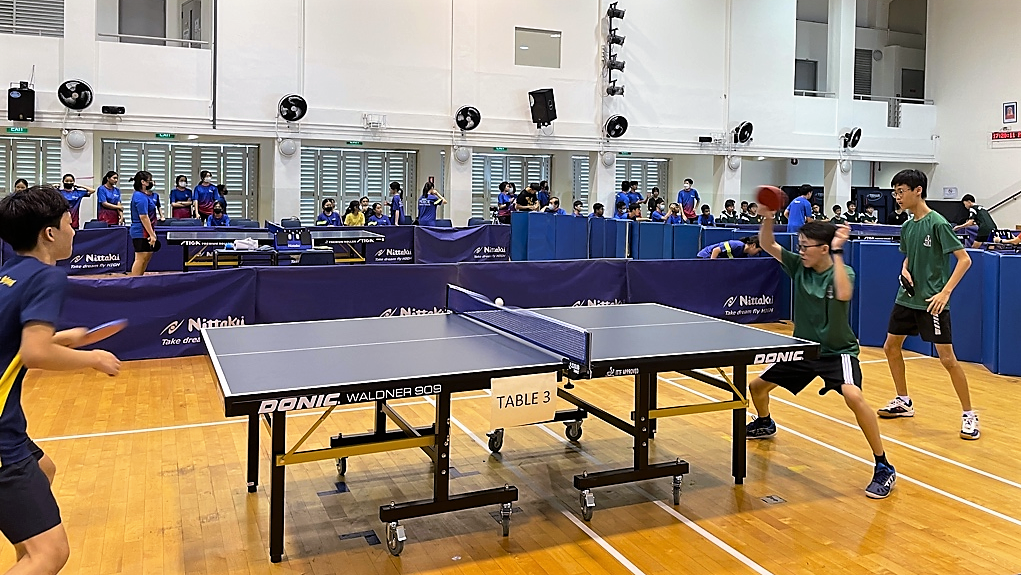 A student in green plays table tennis against an opponent in blue, with spectators and other matches visible in the background.