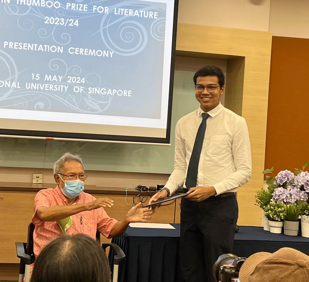 A young man in a white shirt and tie receives a certificate at the Thumboo Prize for Literature 2023/24 ceremony at NUS.