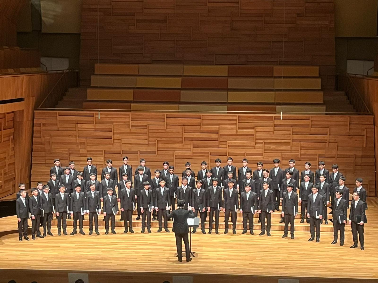 A conductor leads a large boys choir in black suits on a wooden concert hall stage.