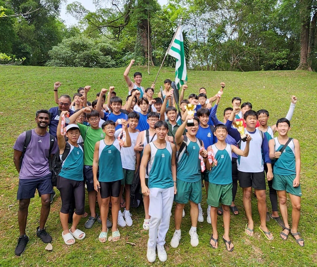 A large group of young male athletes in teal uniforms celebrate outdoors on grass, raising fists and holding a trophy and flag.