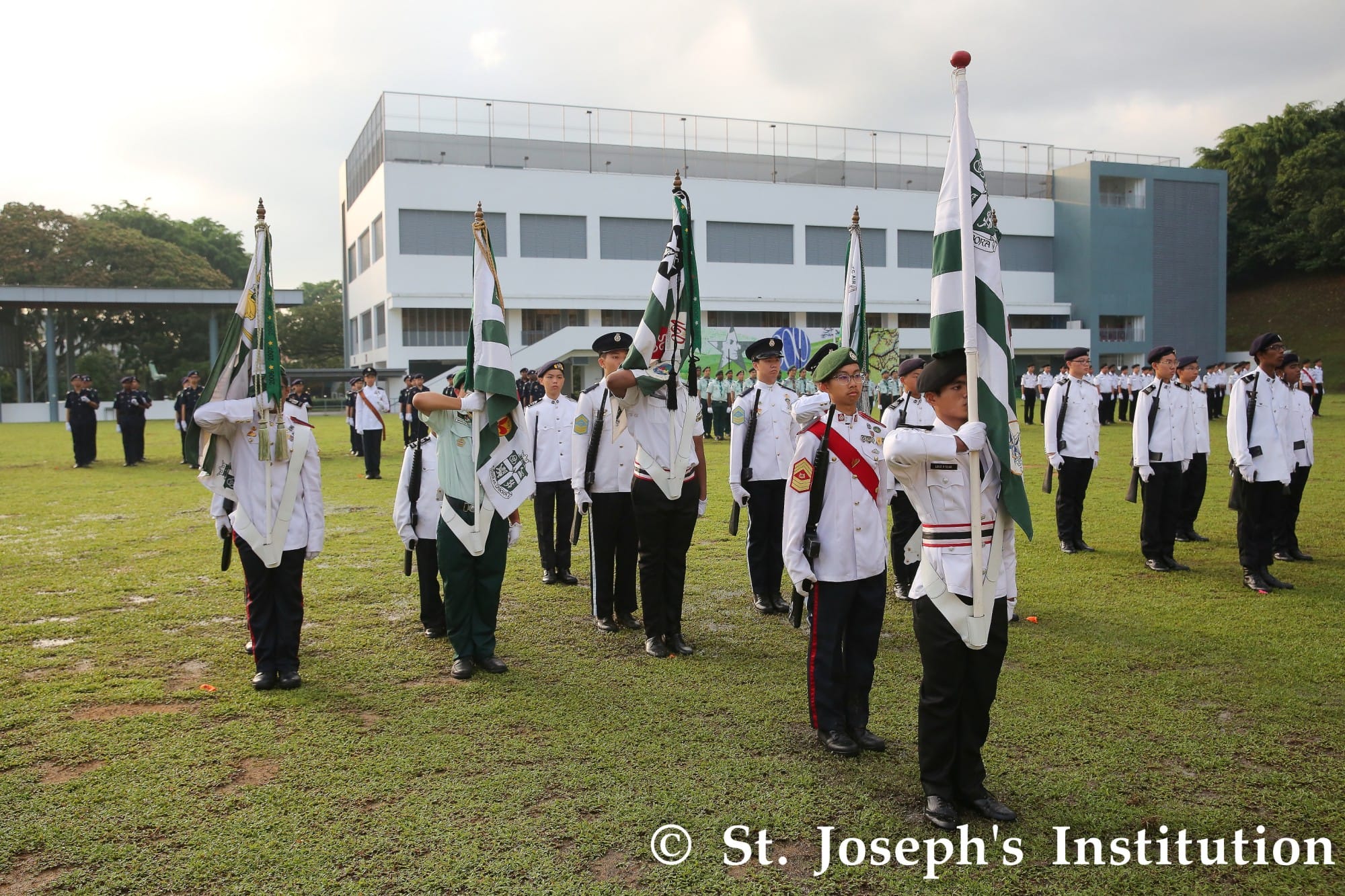 Students in white uniforms carry green and white flags during a formal parade on a school field.