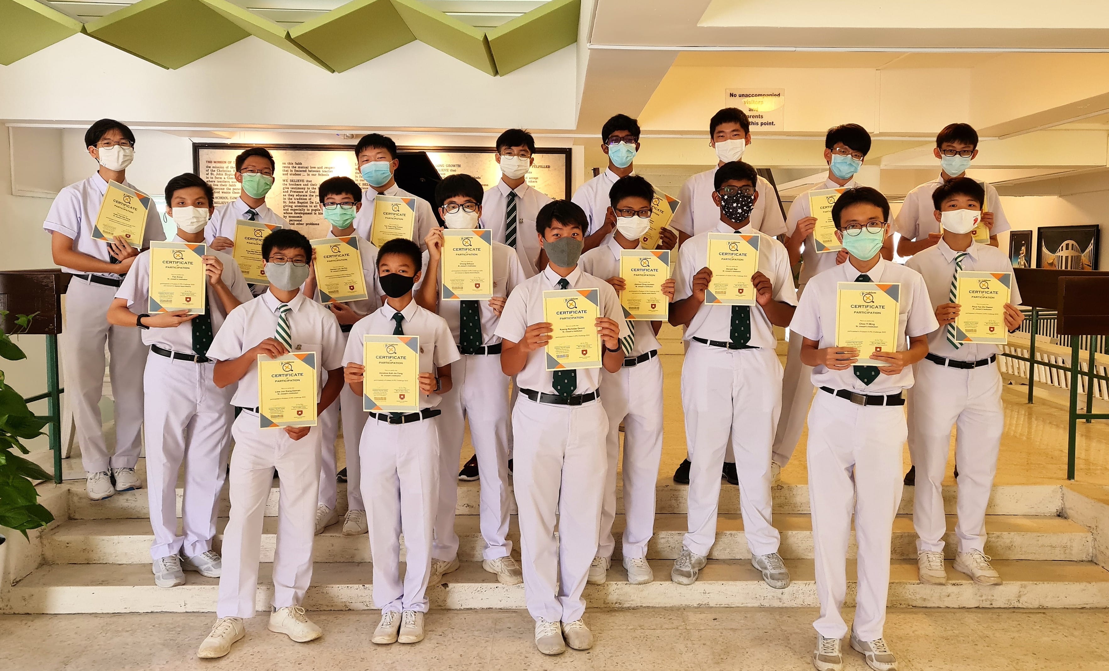 A group of masked male students in white uniforms stand on steps holding participation certificates.