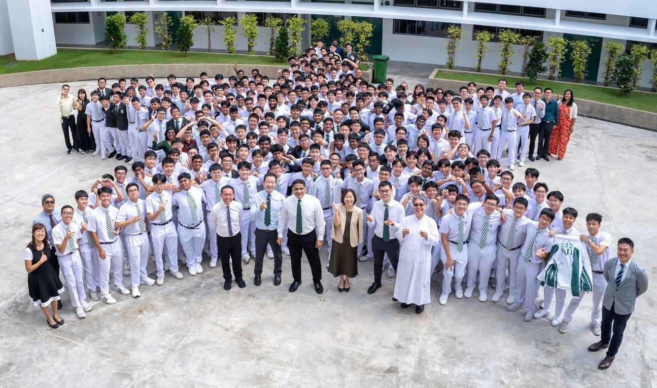 Large group of students in white uniforms with green striped ties pose with teachers in a school courtyard.